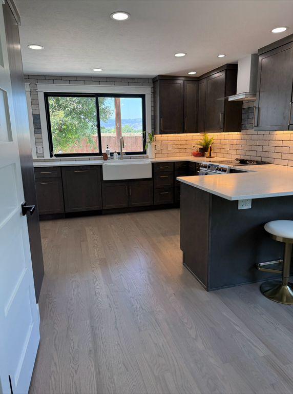 Kitchen with dark cabinets, white farmhouse sink, large window, brick backsplash, wooden floor, and an island with a white countertop and a barstool.