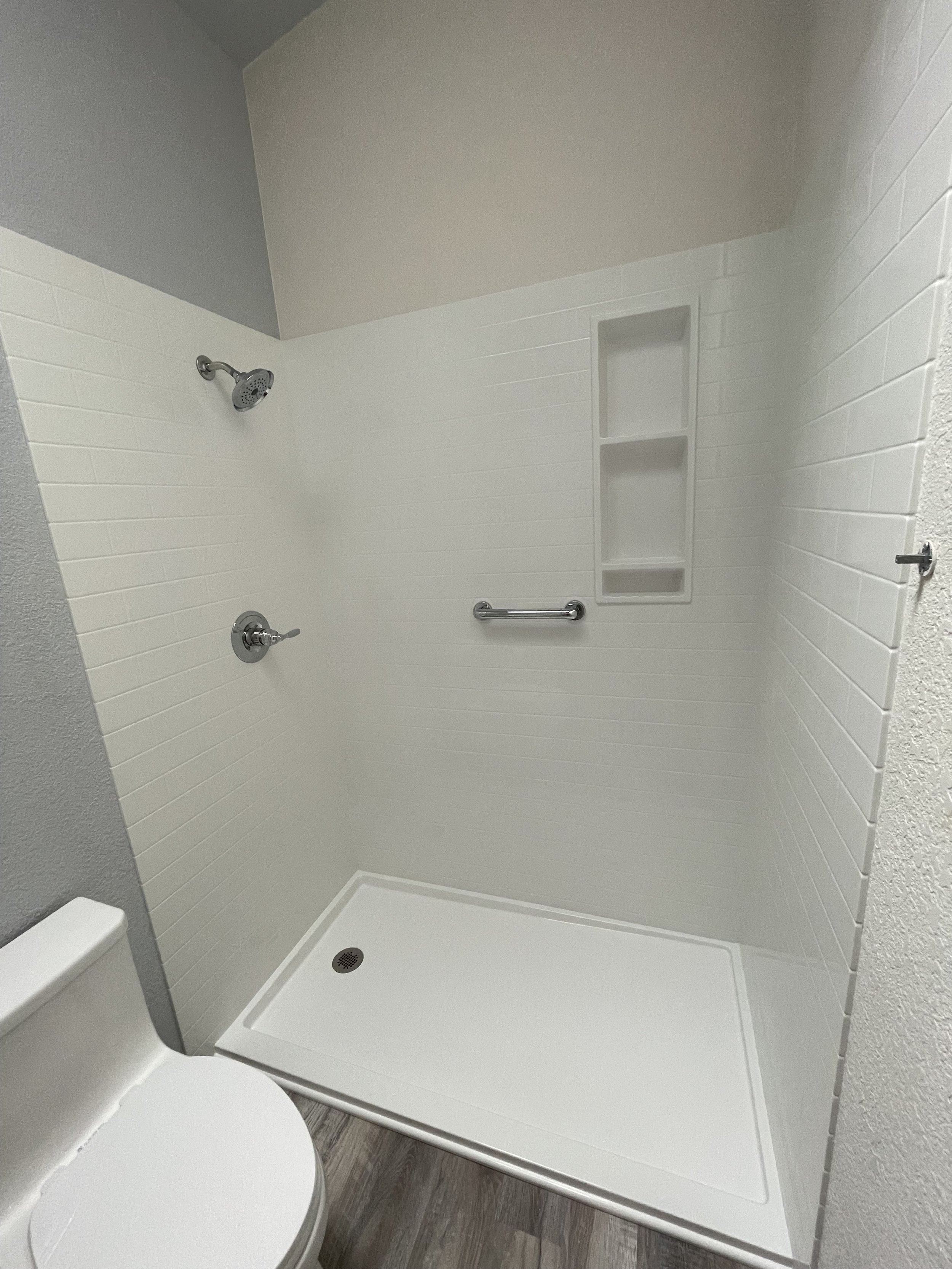 Empty walk-in shower with white subway tiles, a silver grab bar, built-in shelves, a silver showerhead, and a drain in the floor. Part of a nearby white toilet is visible.