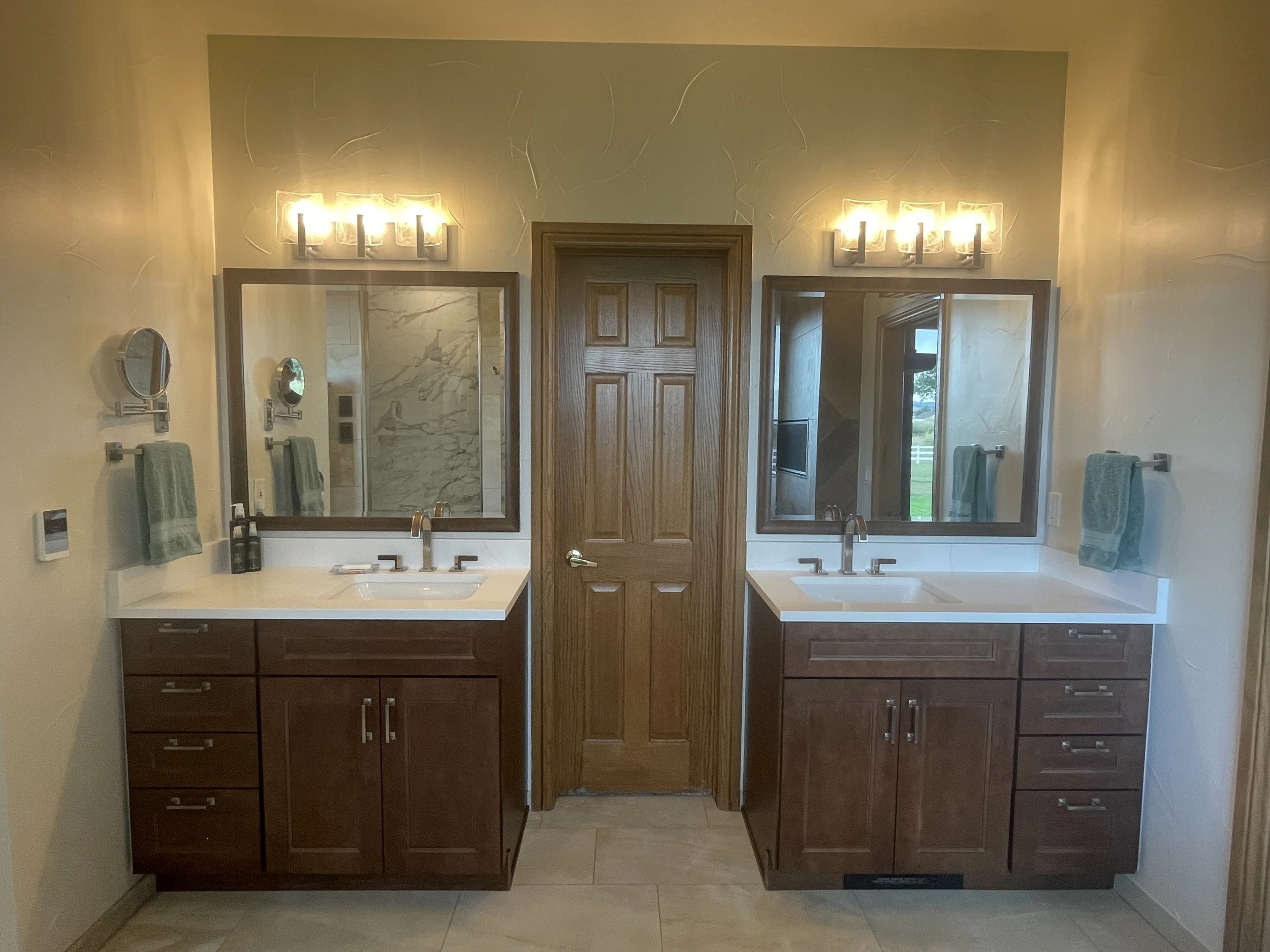 A bathroom with two sinks, two mirrors, and wooden cabinets underneath each sink. There is a wooden door in the center and towel racks with green towels on each side. The lighting fixture above the mirrors has four bulbs.