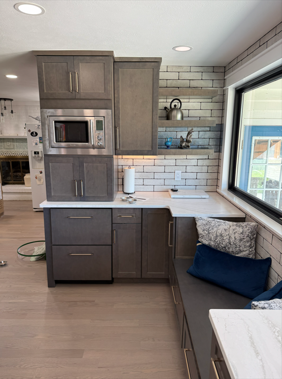 Kitchen corner with gray cabinetry, a microwave, a paper towel holder, a window seat with pillows, and a brick wall with a shelf holding a kettle and decorative items.