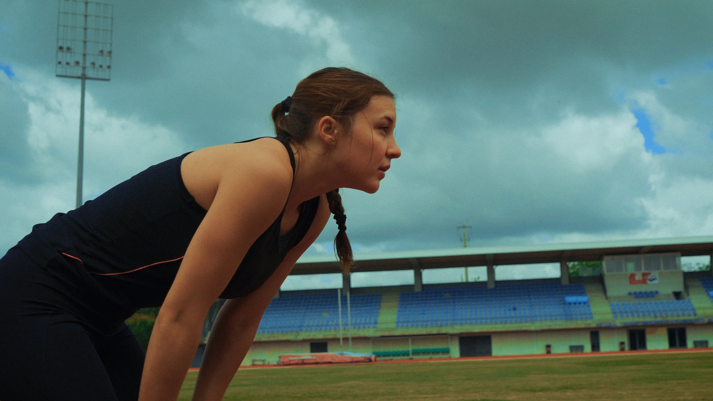 Atleta feminina agachada no estádio, se preparando para correr, sob céu nublado.