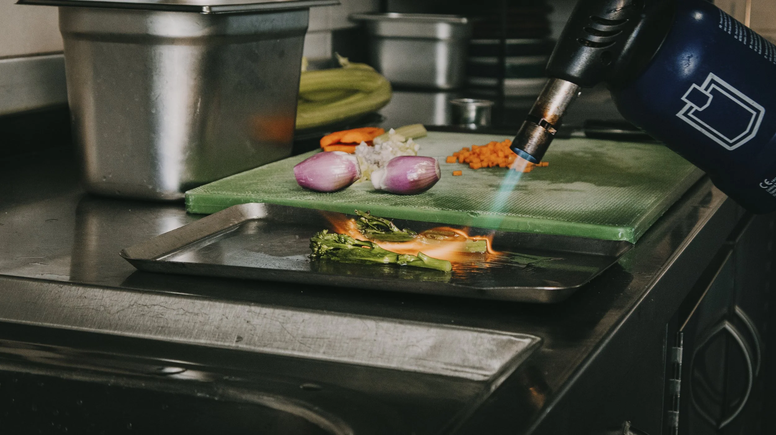 Cozinha com fogo soprando em uma panela com legumes, incluindo alhos, cenoura e folhas verdes, ao lado de uma tábua de cortar com mais legumes, em uma cozinha profissional.