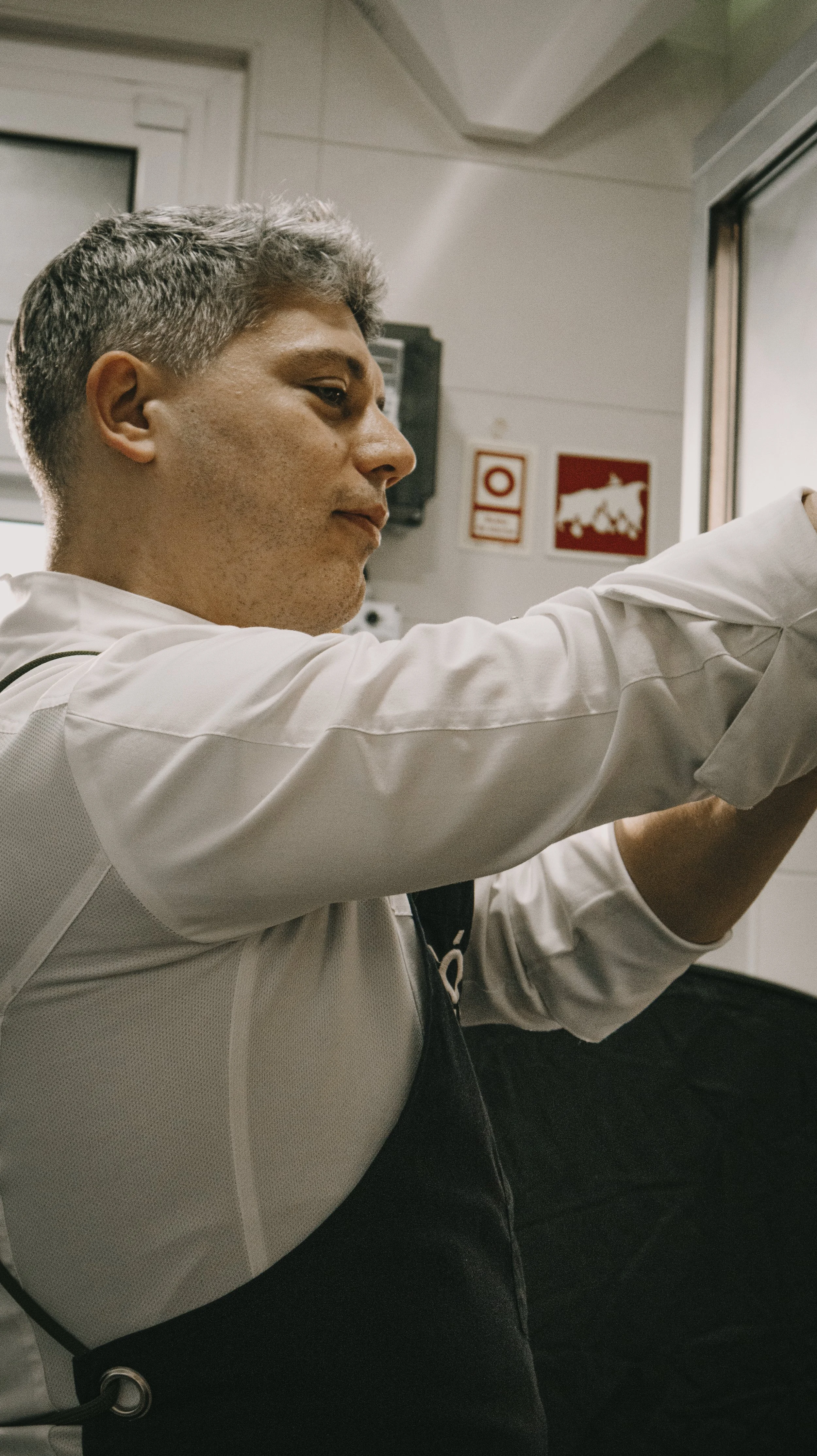 Homem de cabelo grisalho em uniforme de trabalho ajustando algo na parede, dentro de uma sala com avisos de emergência ao fundo.