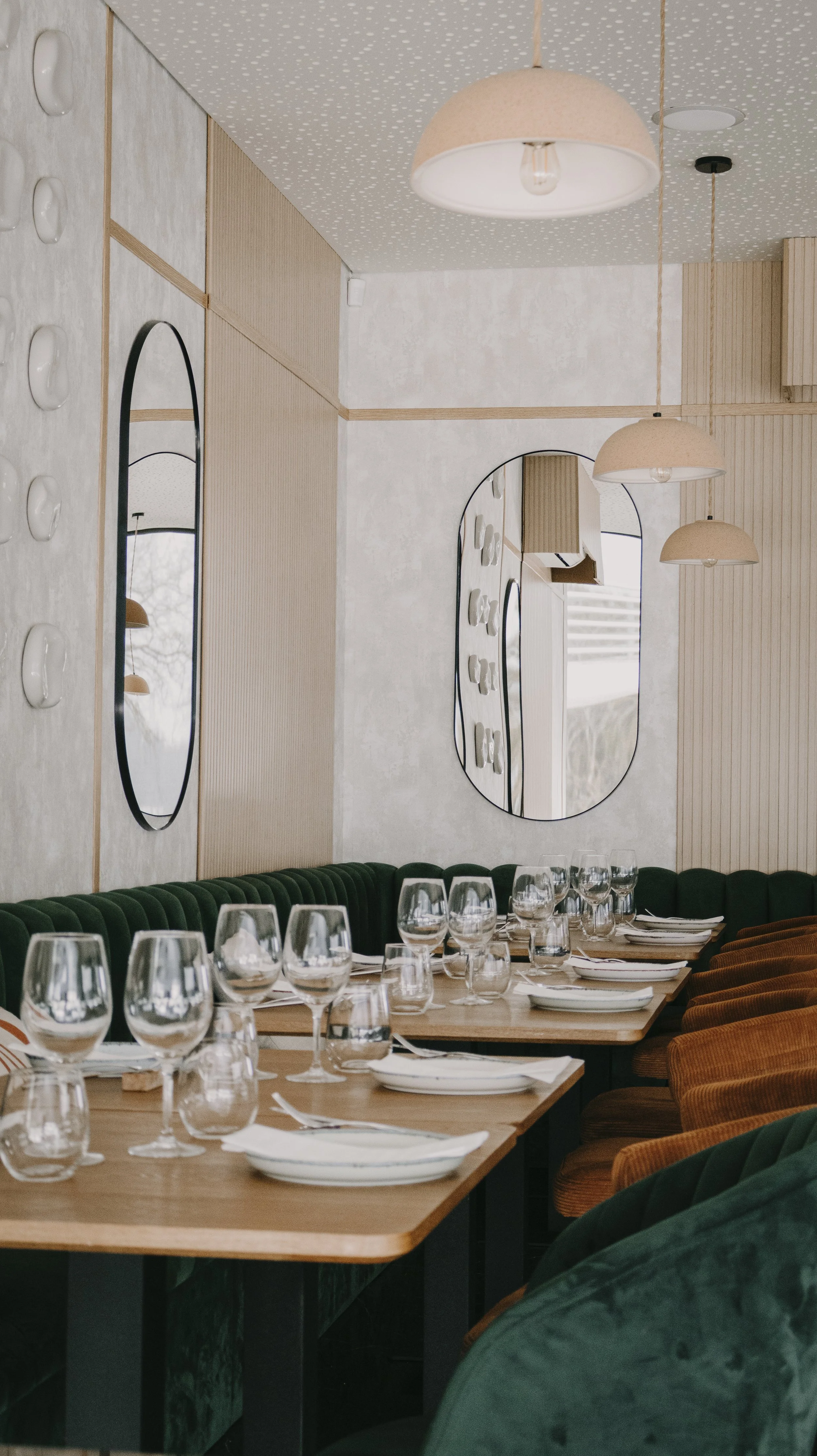 Sala de jantar com mesas arrumadas, taças de vinho, pratos brancos, cadeiras de veludo verde e laranja, espelhos retangulares e redondos nas paredes, luminárias suspensas de cor clara e paredes decoradas com painéis de madeira clara e detalhes branco