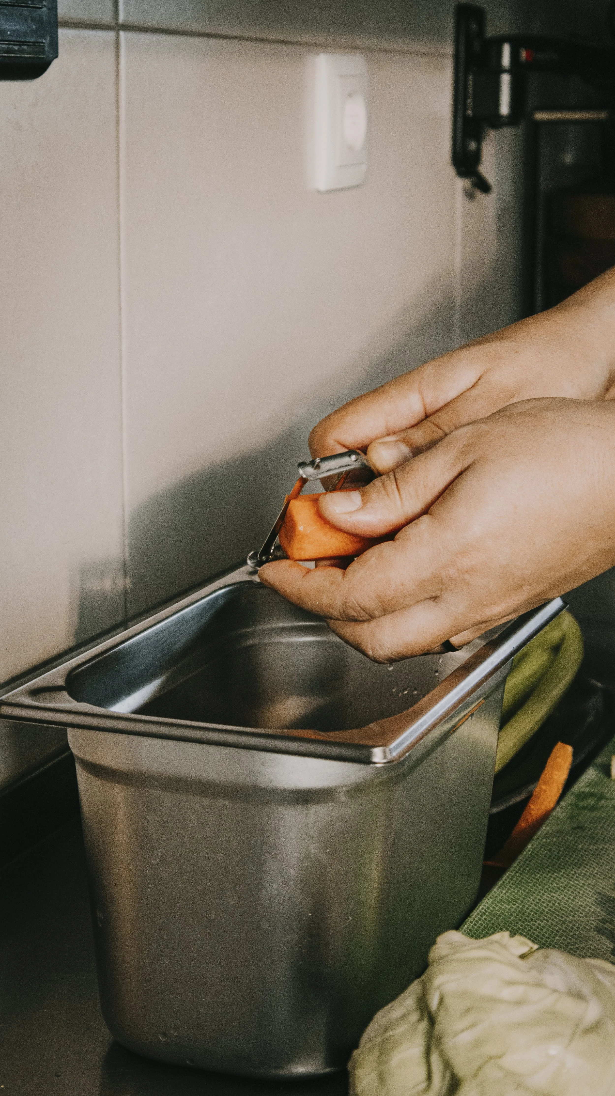 Pessoa usando um descascador para descascar uma cenoura sobre uma tigela de aço inoxidável.