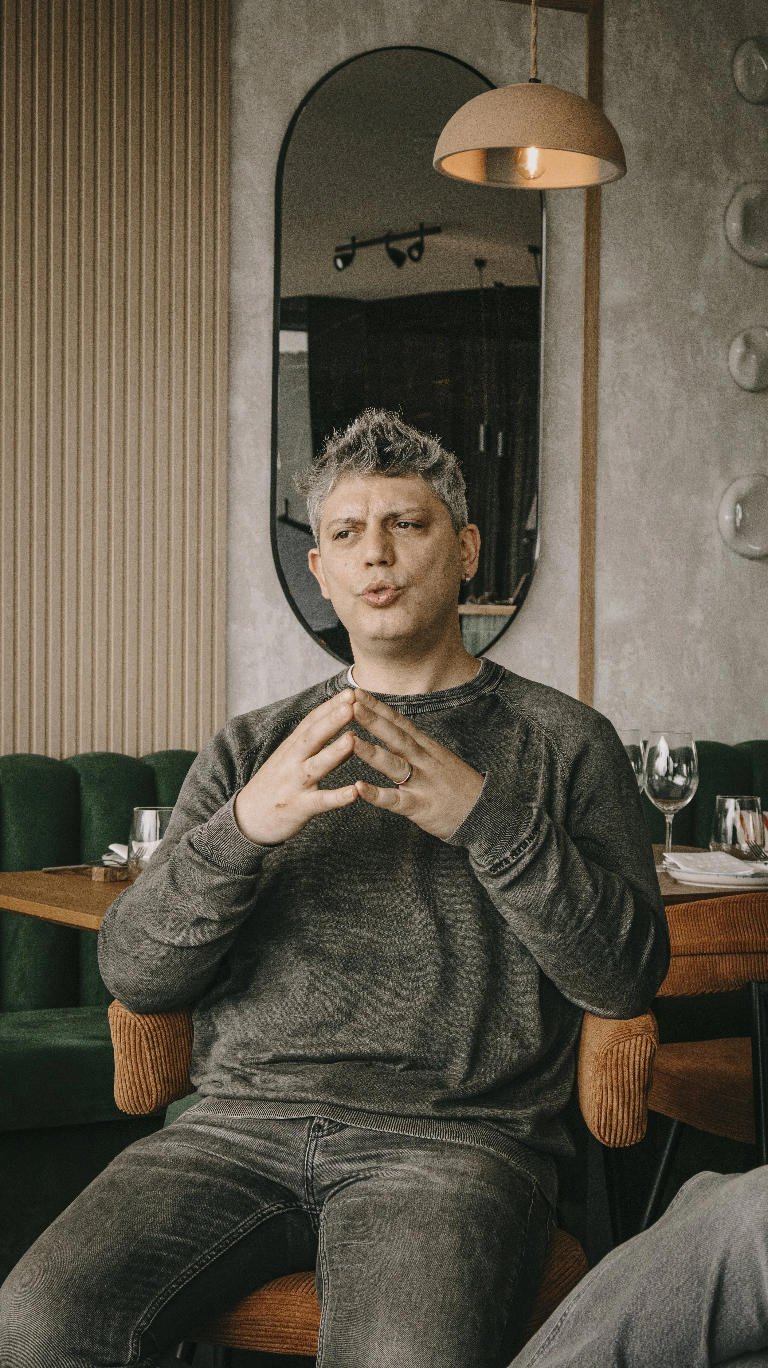 Homem sentado em um restaurante com expressão pensativa, mãos juntas na frente do rosto, ao lado de uma mesa com taças de vinho.