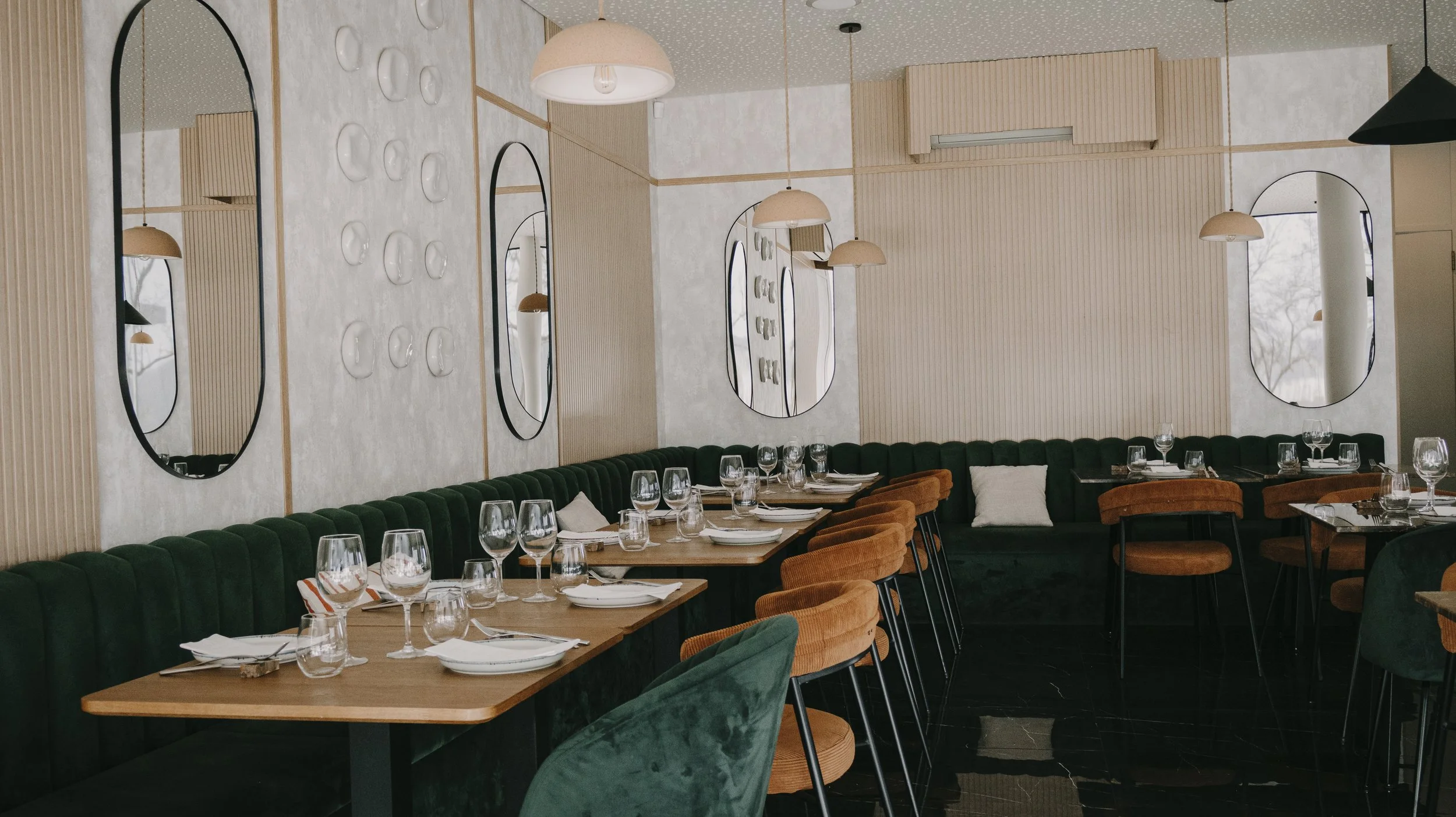 Interior de restaurante com paredes decoradas, espelhos ovalados, iluminação amarelada, mesas com taças e pratos, cadeiras de veludo verde e estofados marrons.