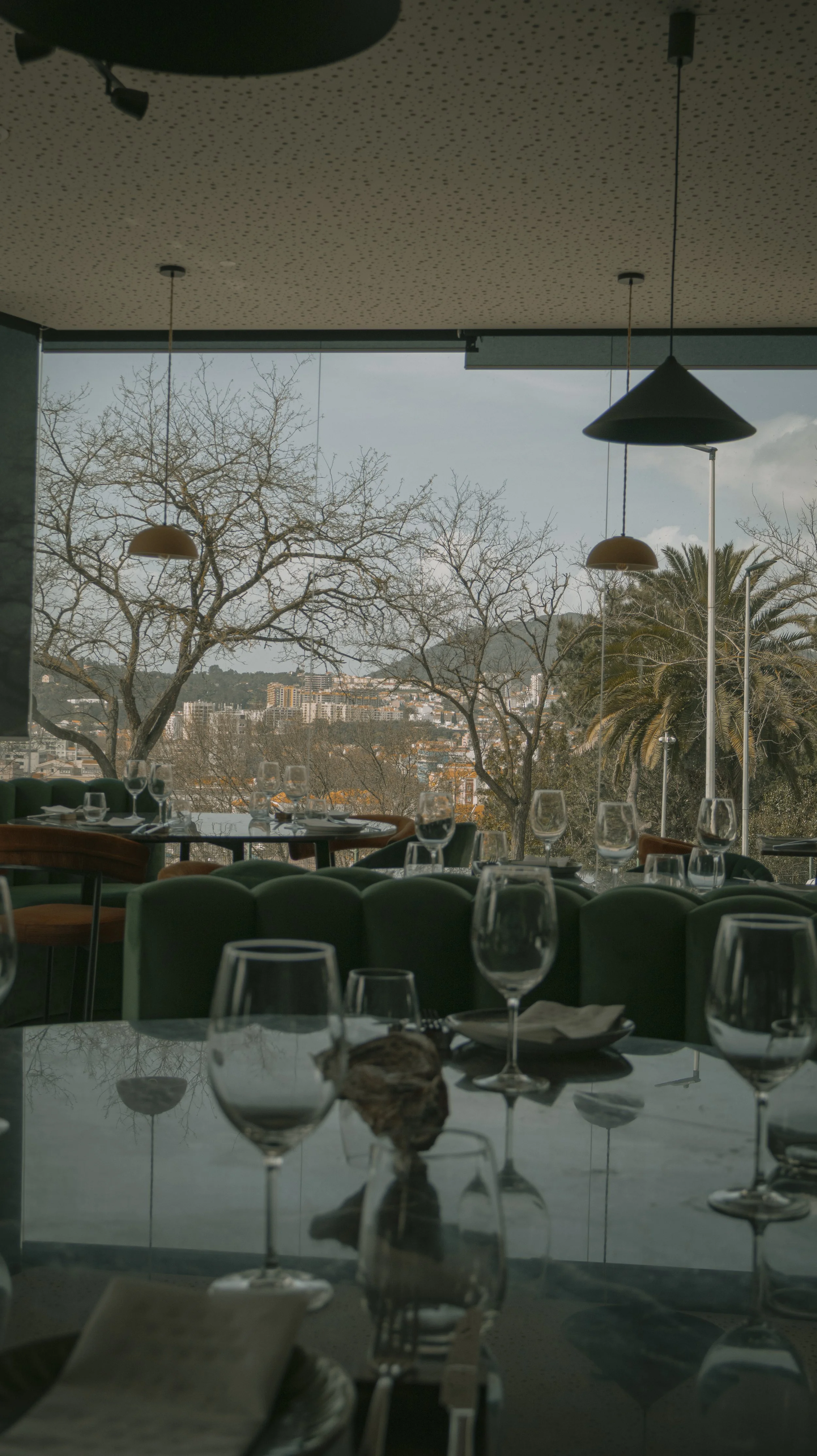 Interior de restaurante com mesas preparadas, copos de vinho e vista para árvores e cidade ao fundo