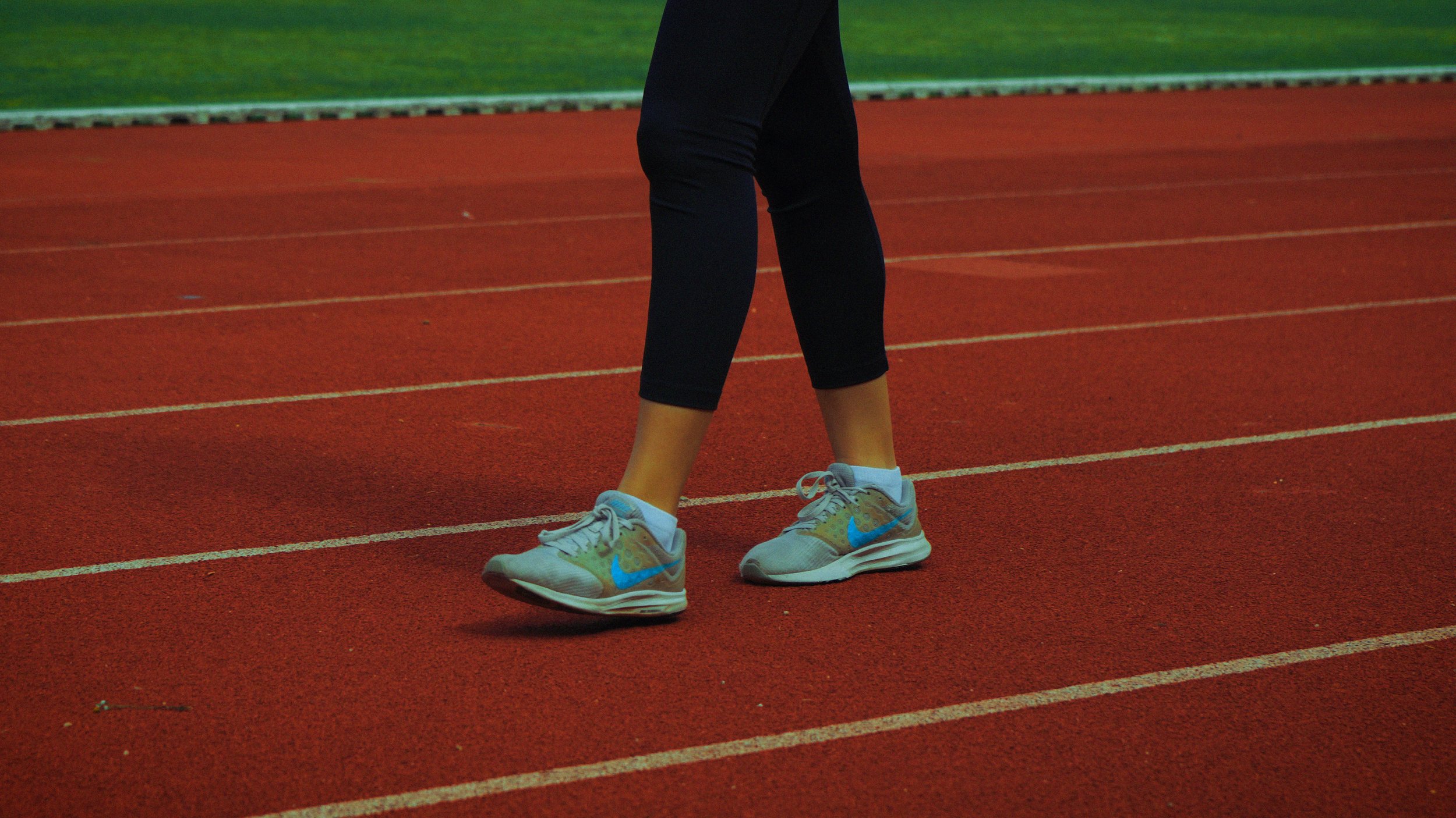 Pista de corrida vermelha com linhas brancas, vista de pernas de pessoa com calçados esportivos brancos e detalhes azuis e preta, de roupas pretas, em uma pista de atletismo.