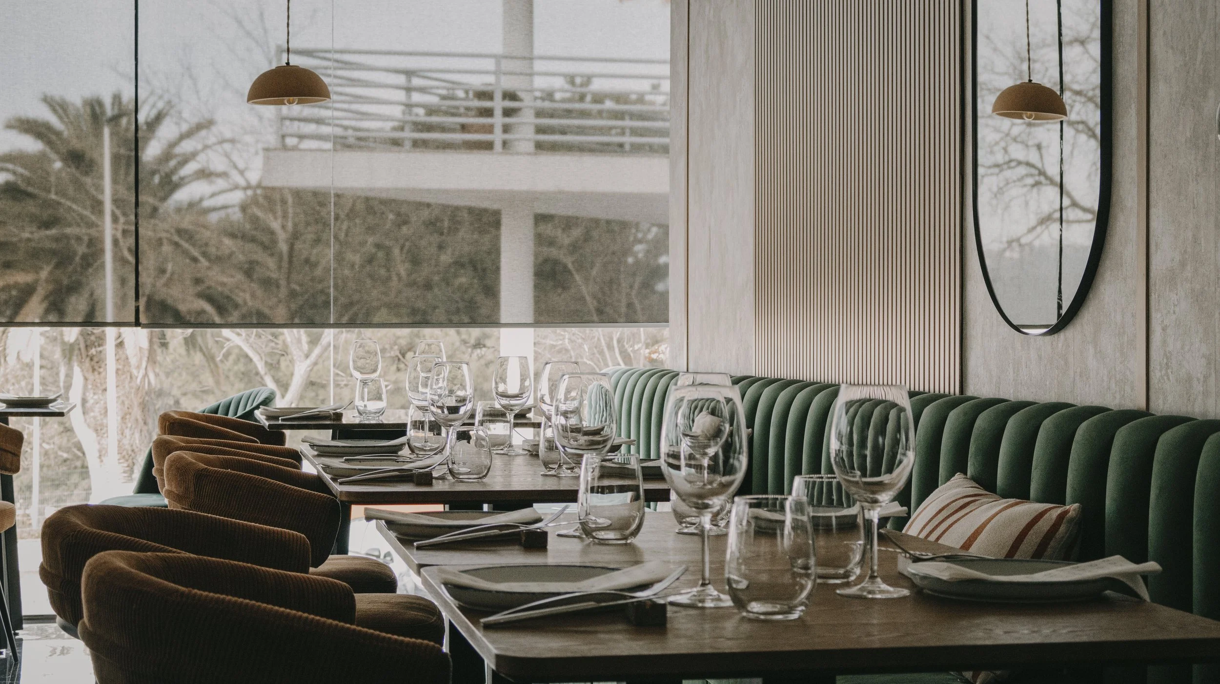 Mesa de jantar em restaurante com taças, pratos e talheres, cadeiras de veludo coloridas e janela grande com vista para árvores.