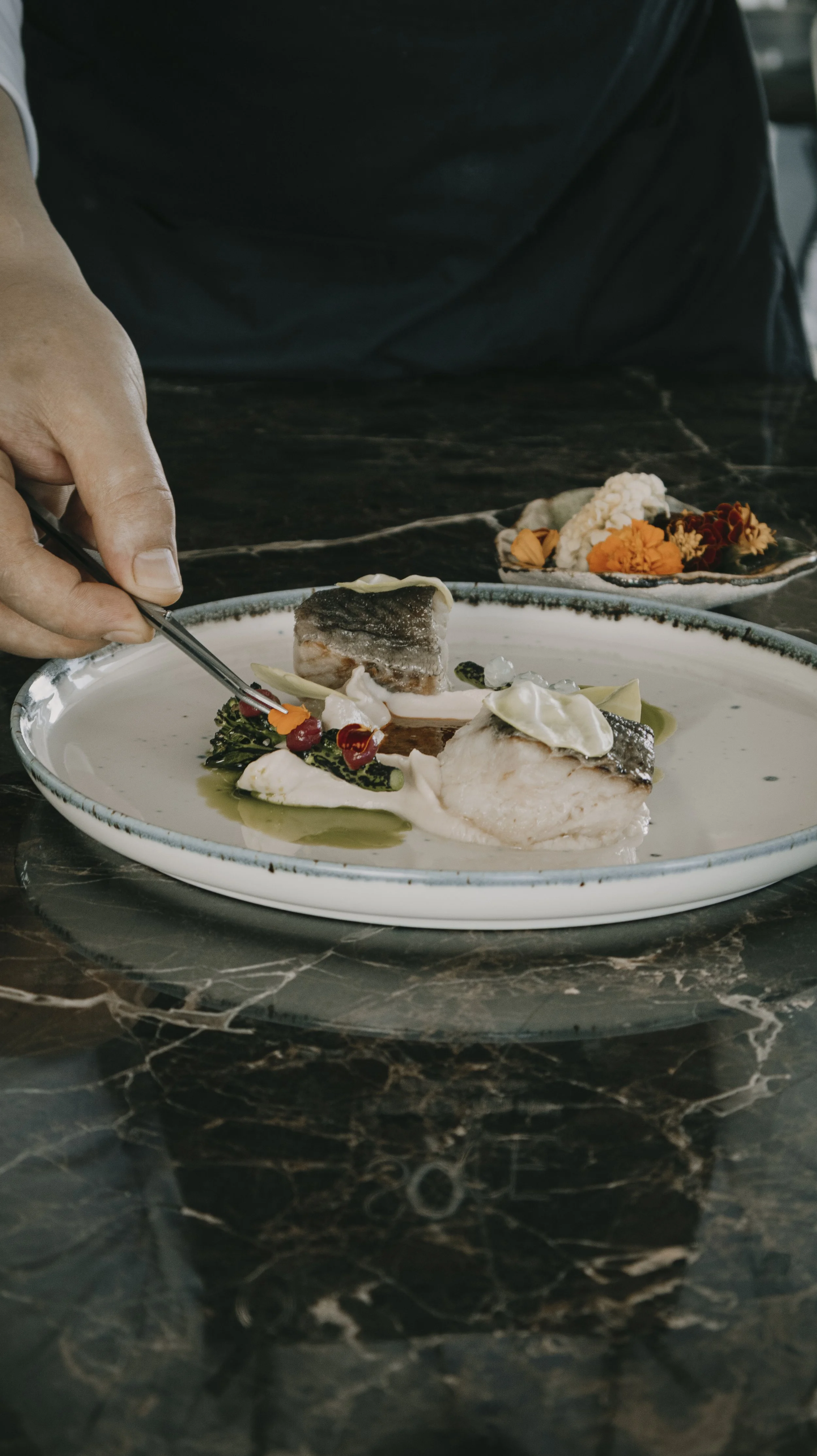 Prato com peixe grelhado, folhas verdes, molho branco e decorativos com flores comestíveis, em um restaurante. Ao fundo, prato menor com flores.