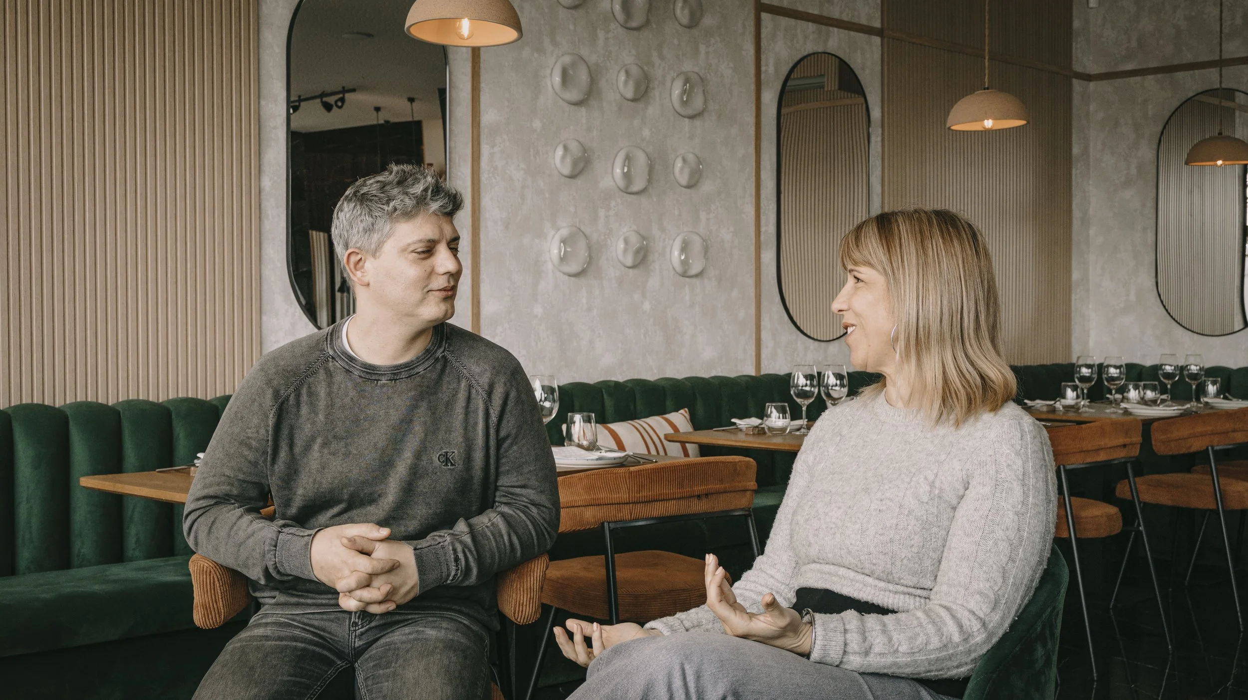Homens e mulheres conversando em um restaurante com decoração moderna, mesas com talheres e taças de vinho ao fundo.