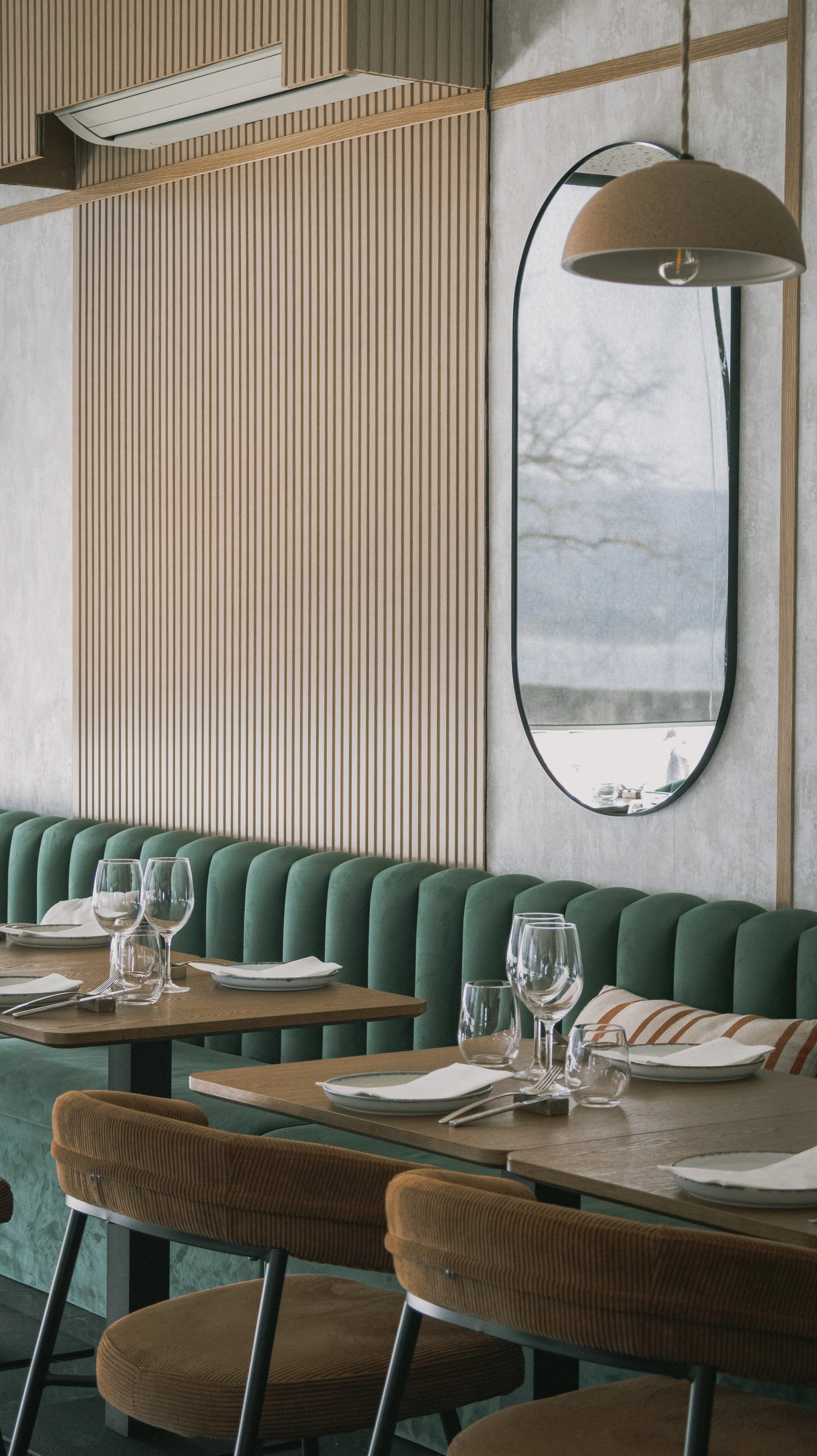 Interior de restaurante com mesa posta, copos e pratos, cadeiras de veludo verde e poltrona marrom, parede de painéis de madeira clara, espelho oval e luminária pendente