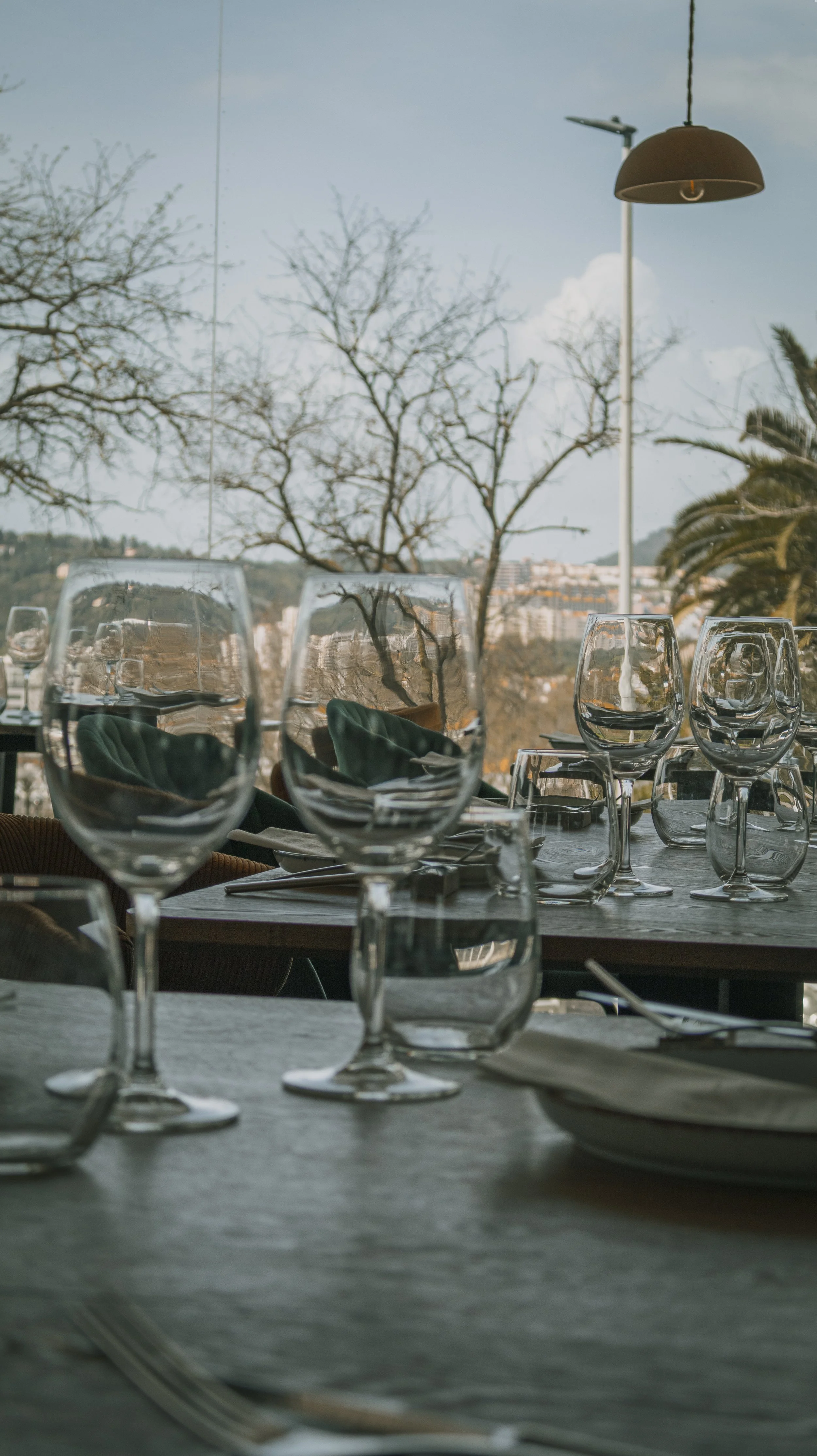 Mesa de restaurante com taças de vidro, pratos e talheres, vista para árvores e cidade ao fundo.