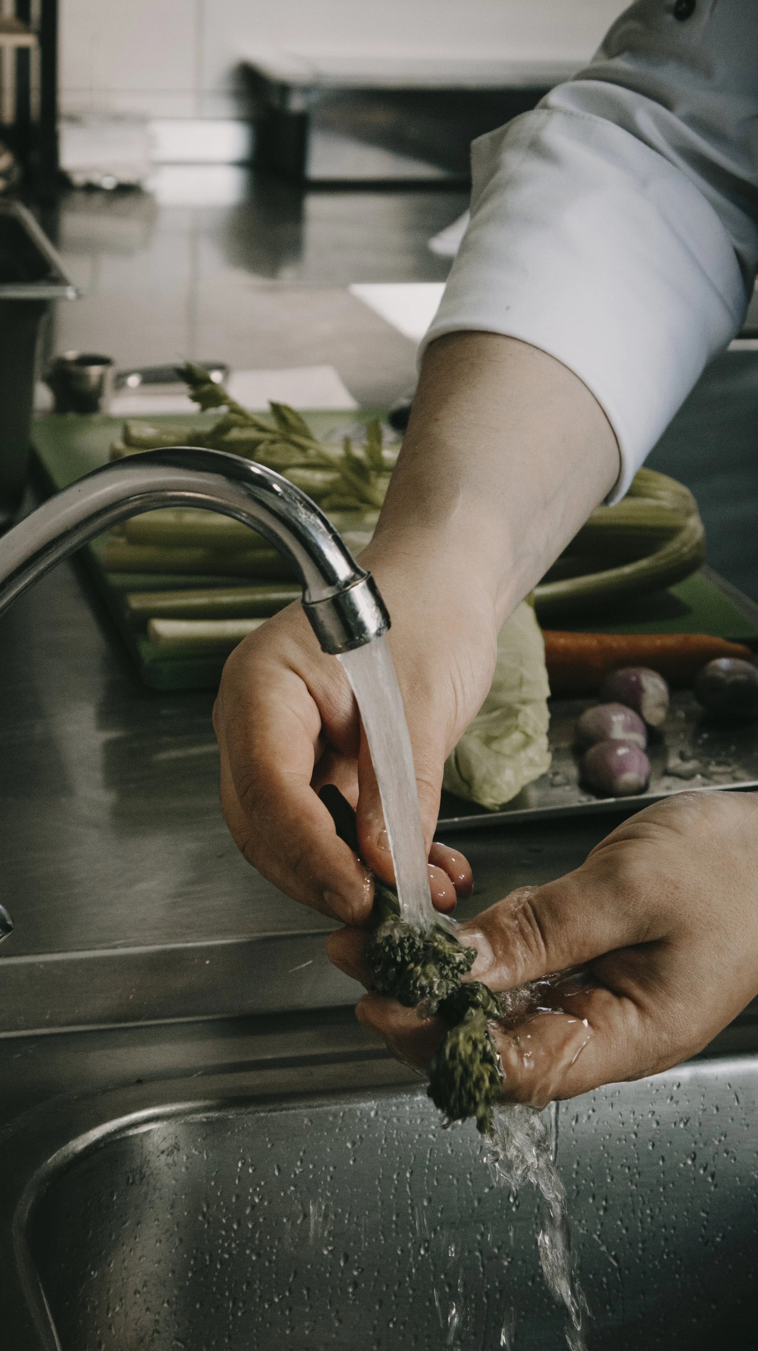 Pessoa lavando brócolis sob torneira na cozinha, com ingredientes e utensílios ao fundo.