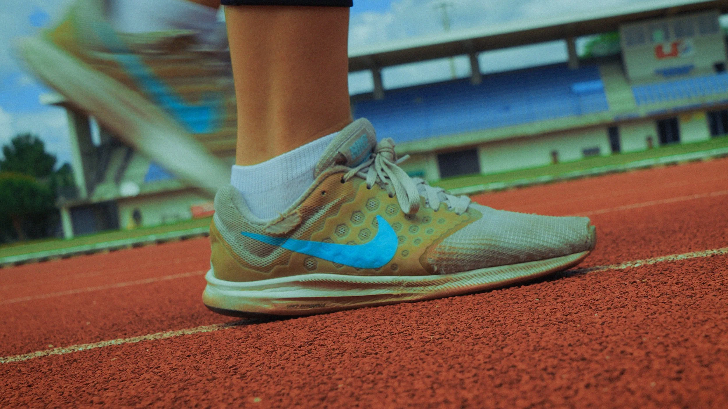 Pé de uma pessoa calçando tênis Nike amarelado e branco em uma pista de atletismo vermelha, com quadra de esportes ao fundo e arquibancadas azuis.