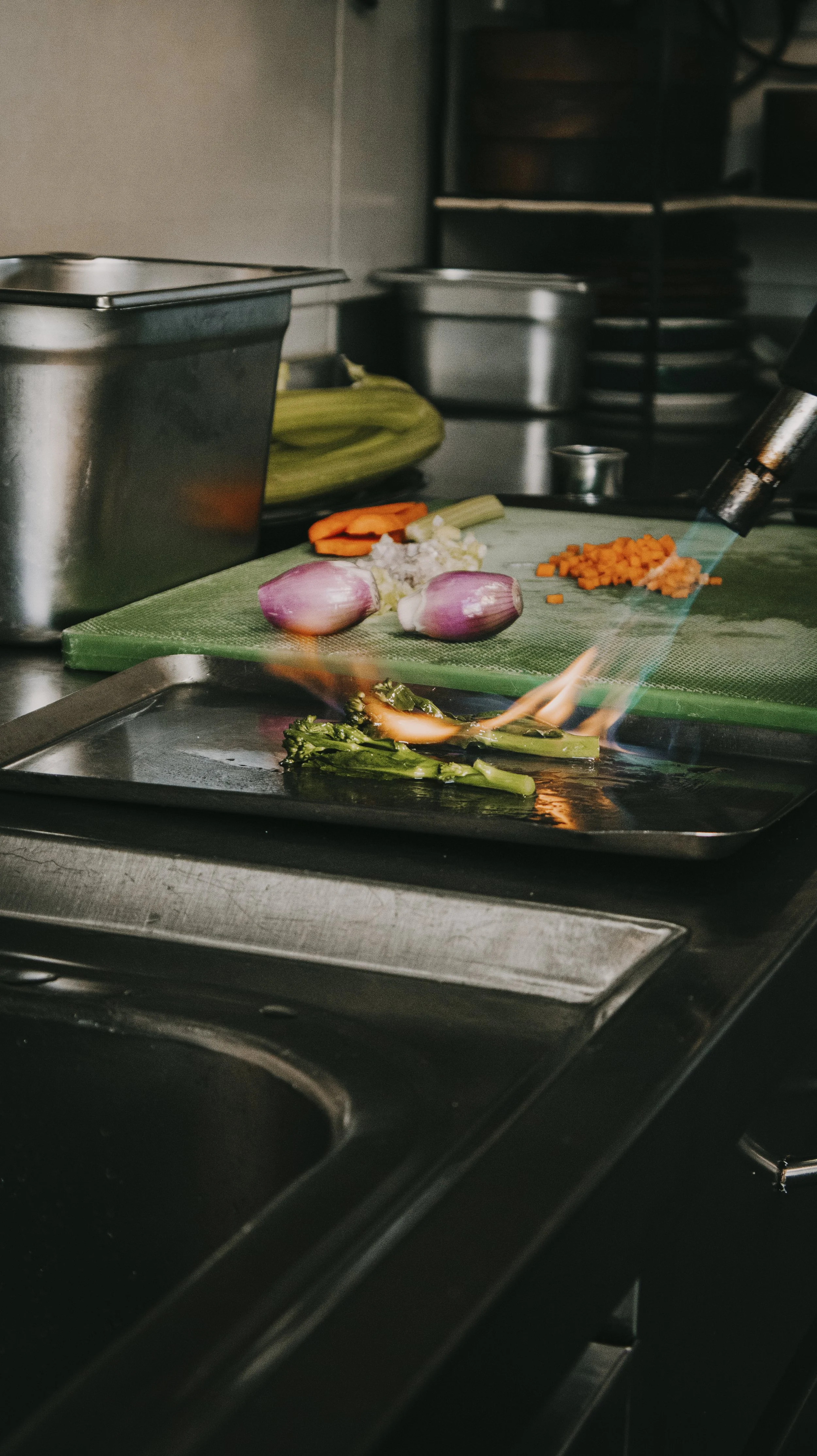 Fogão com vegetais sendo flambados, incluindo brócolis, cebola, cenoura e alho, em cozinha profissional.