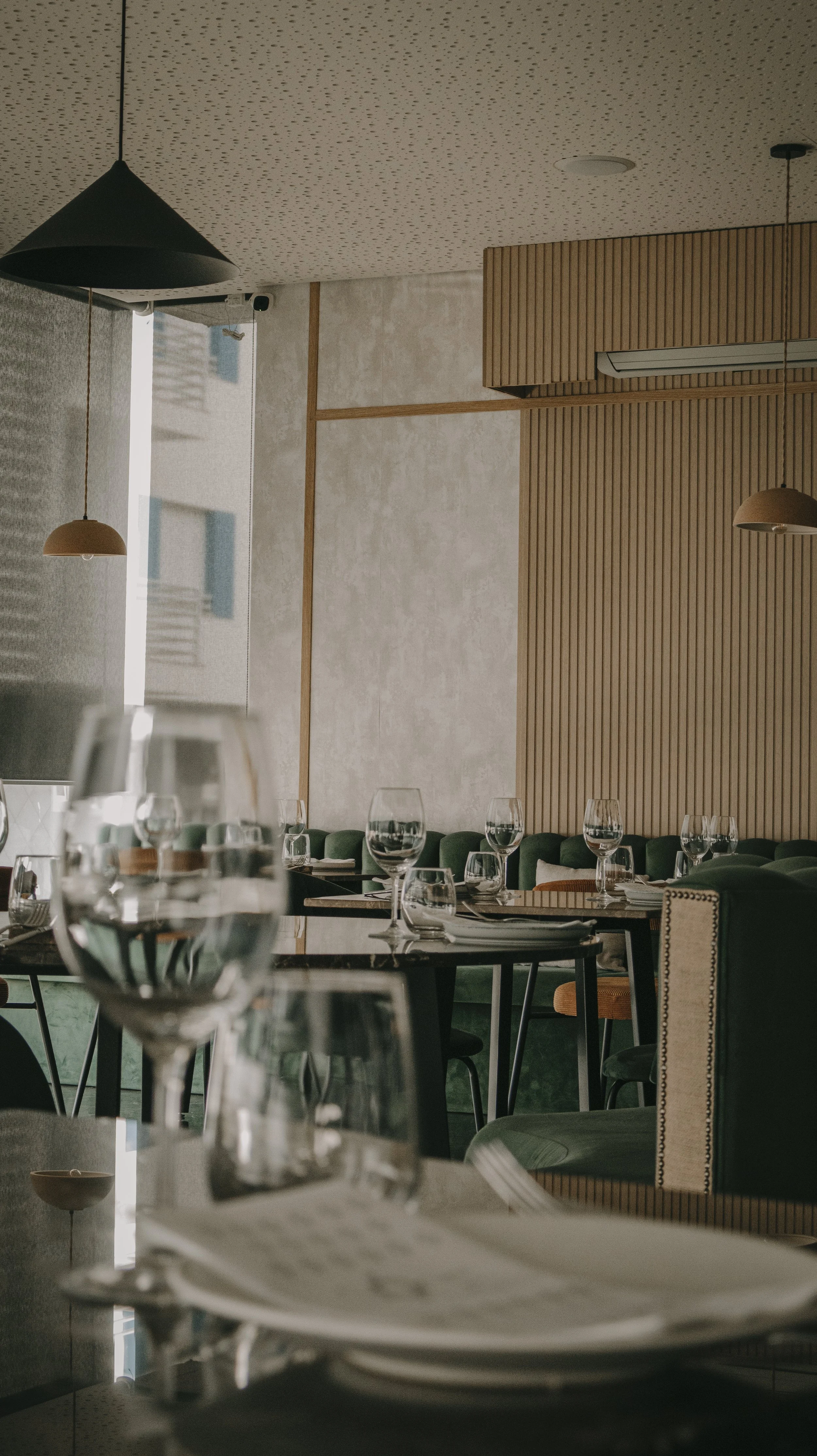 Interior de restaurante elegante com mesas preparadas, copos de água, taças de vinho, pratos e talheres, decorado com paredes de madeira e detalhes modernos, iluminação suave e janelas com cortinas fechadas.