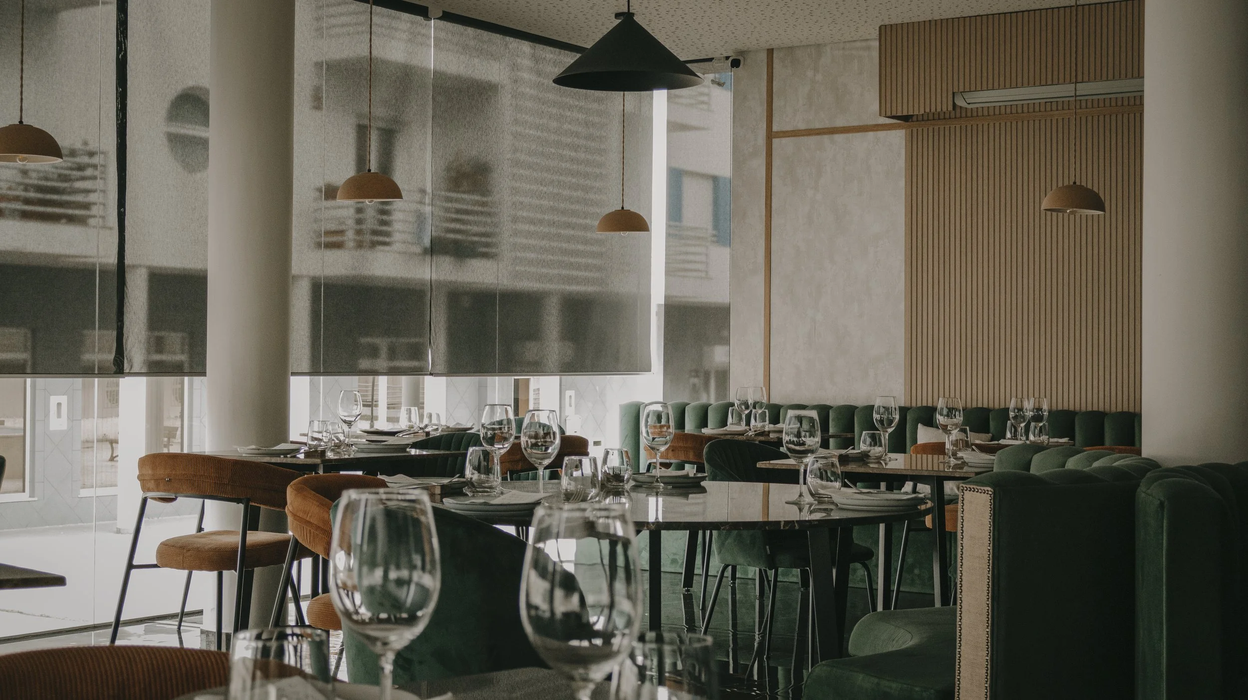 Interior de restaurante elegante com mesas preparadas, taças de vinho e cadeiras verdes e marrons, com iluminação suave e decoração moderna.