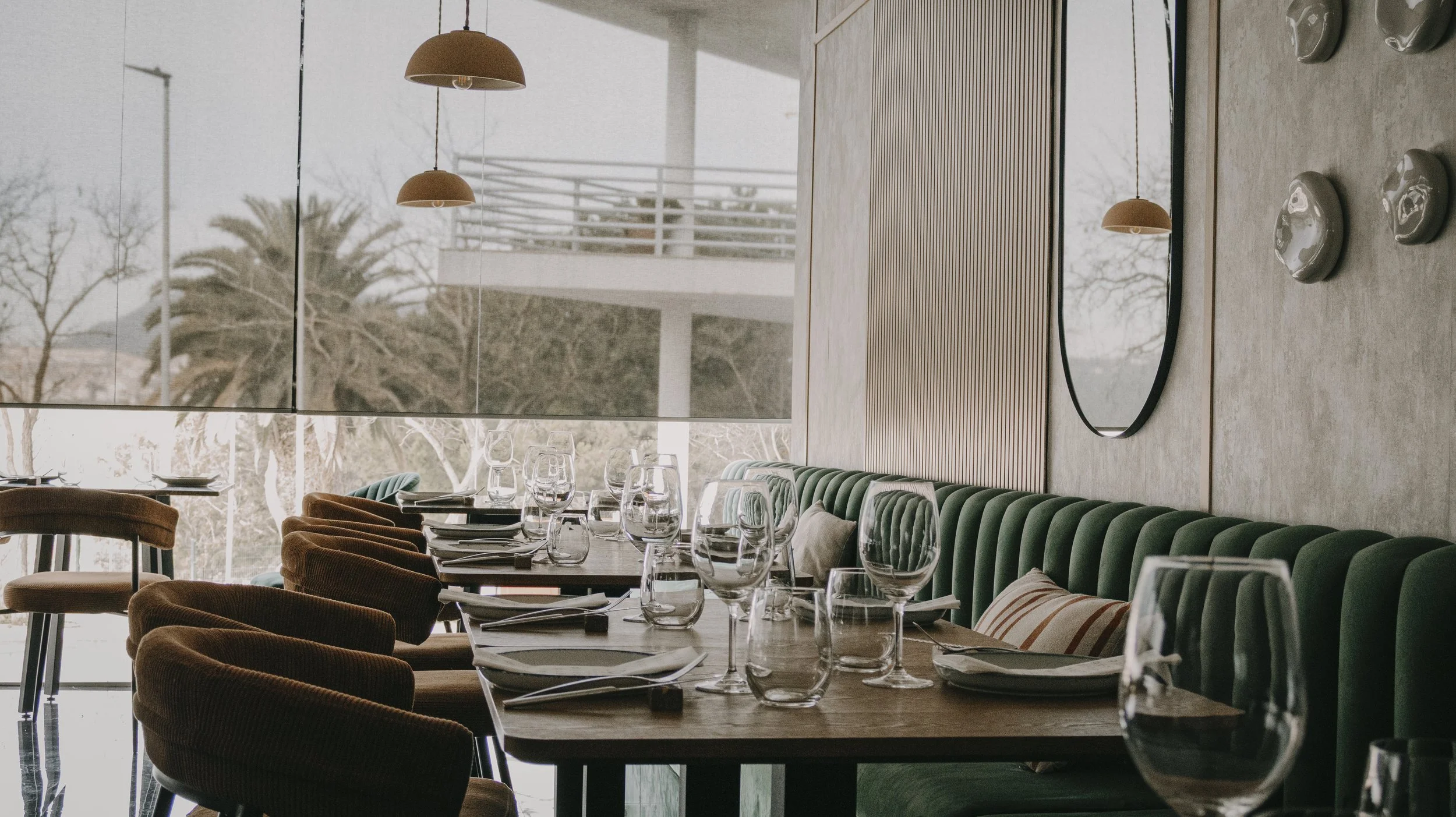 Interior de restaurante elegante com mesa arrumada com pratos, copos de vinho, taças de água e talheres, cadeiras acolchoadas e um banco verde. Grandes janelas oferecem vista de árvores e um prédio ao fundo.
