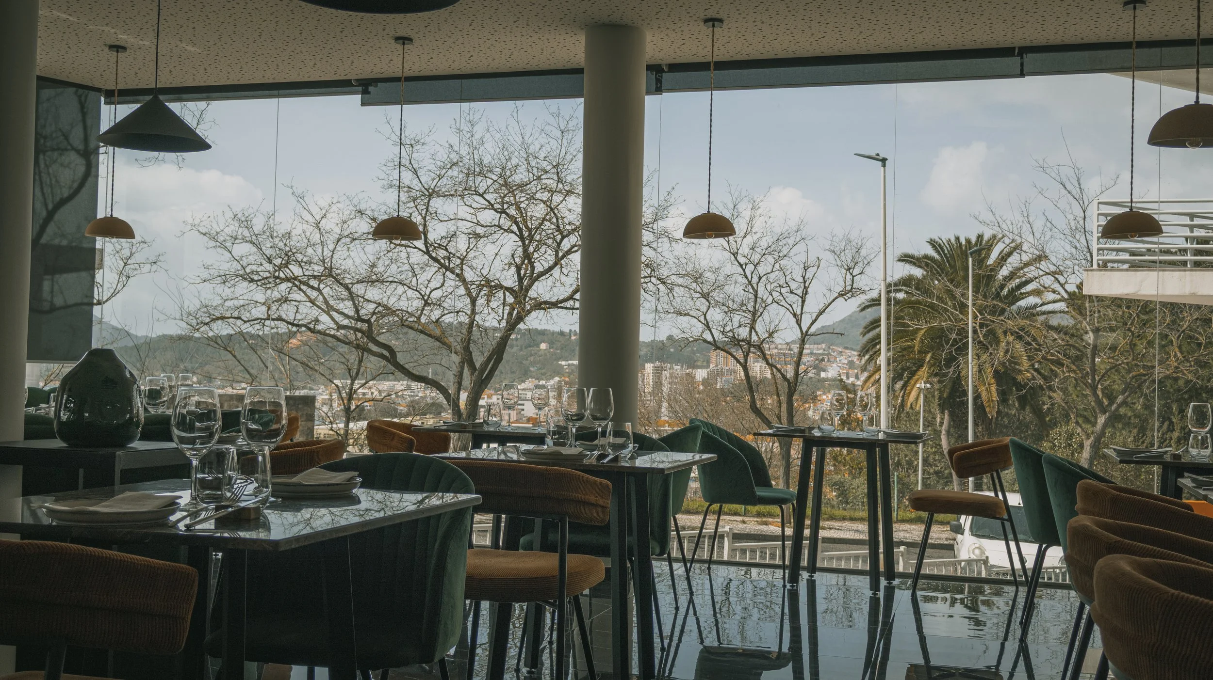 Interior de restaurante com mesas e cadeiras ao lado de janelas oferecendo vista para árvores e cidade ao fundo.