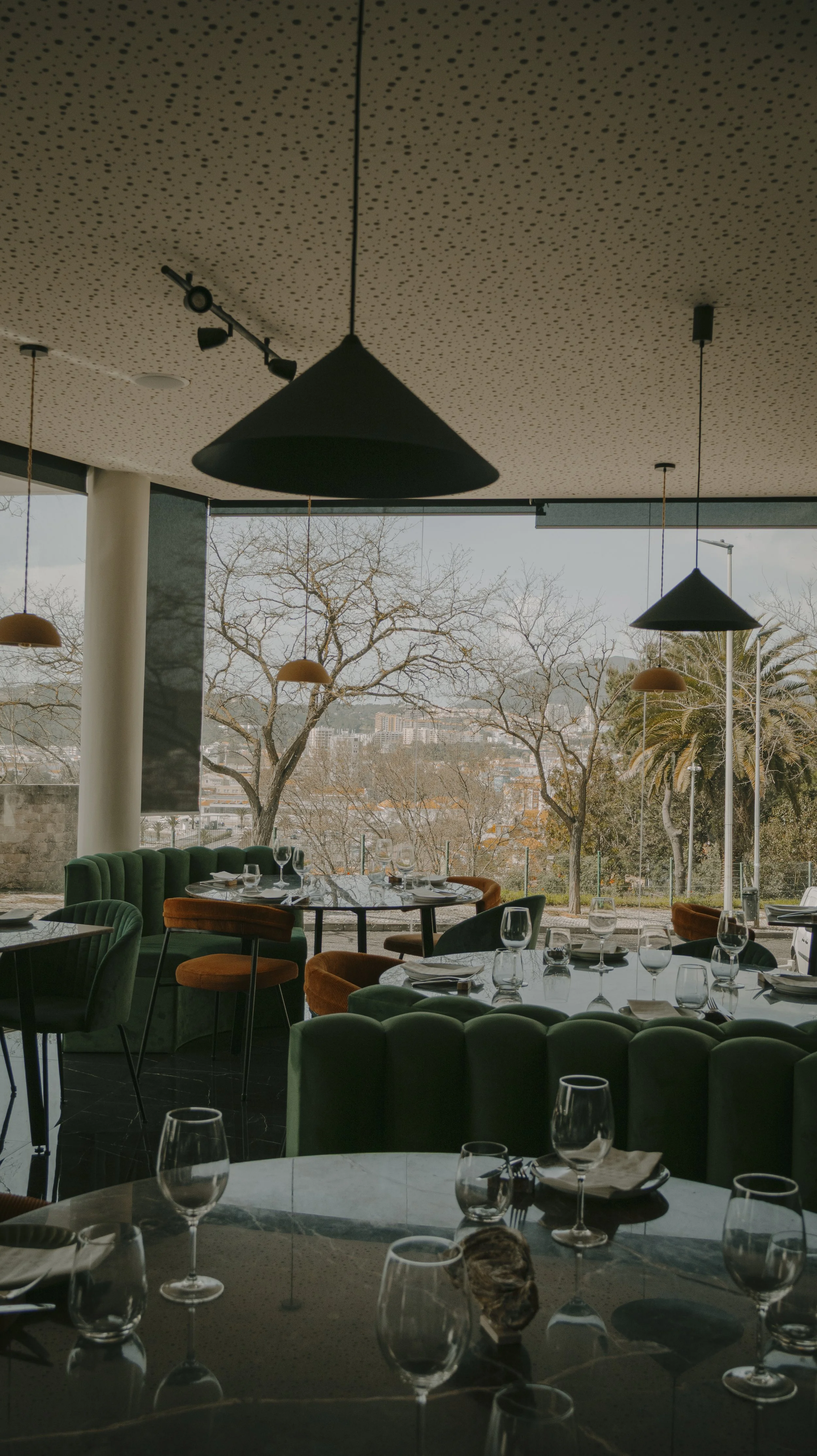 Interior de restaurante com mesas decoradas, cadeiras verdes e laranjas, vista de árvores sem folhas e cidade ao fundo.