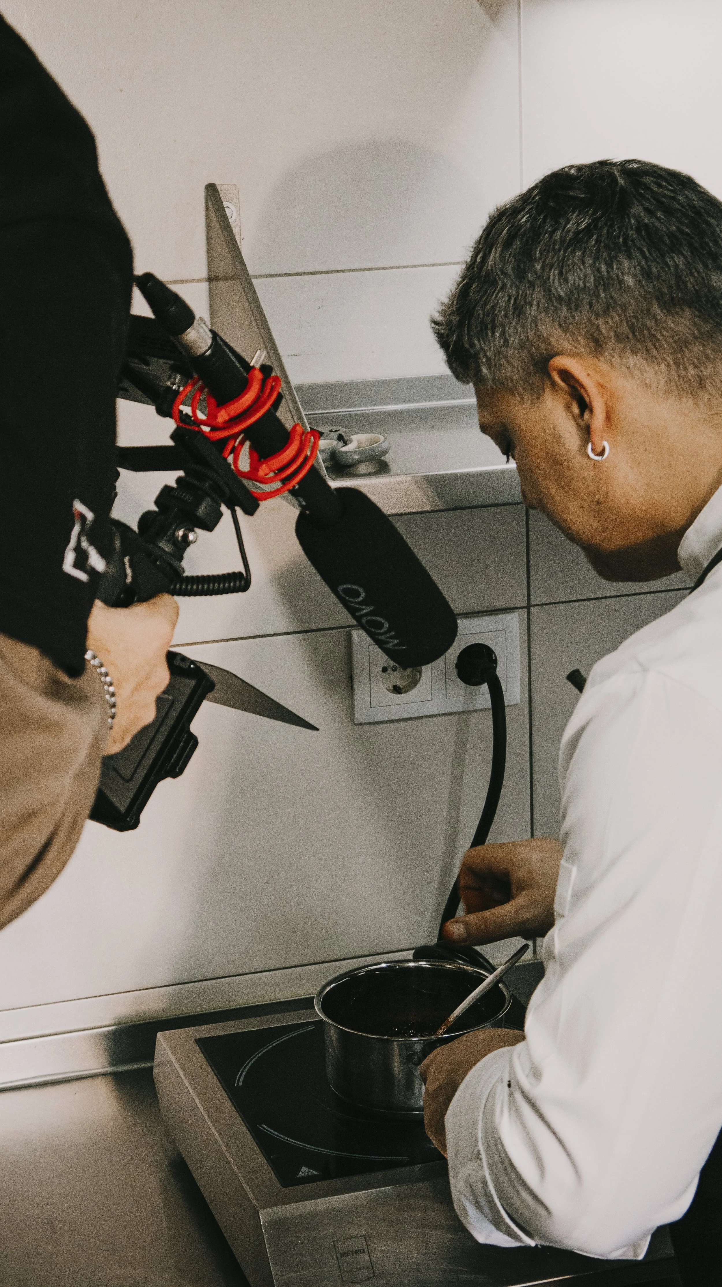 Pessoa cozinhando um molho em uma panela de aço no fogão enquanto outra pessoa grava o procedimento com uma câmera de vídeo com microfone, atrás de uma parede de azulejos na cozinha.