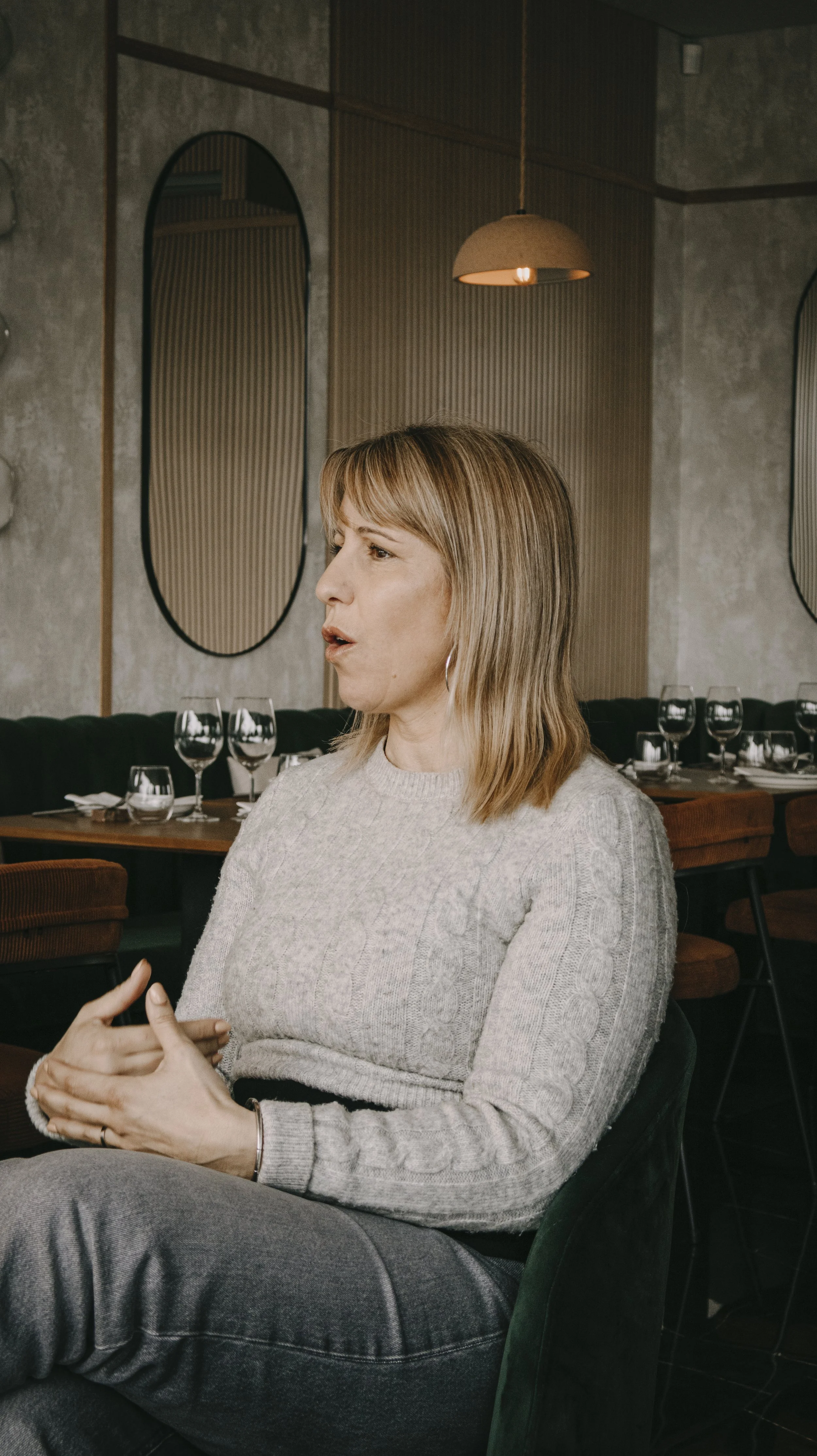 Mulher ruiva, de cabelos médios, sentada em um restaurante, gesticulando enquanto fala. Fundo com mesas e taças de vinho.