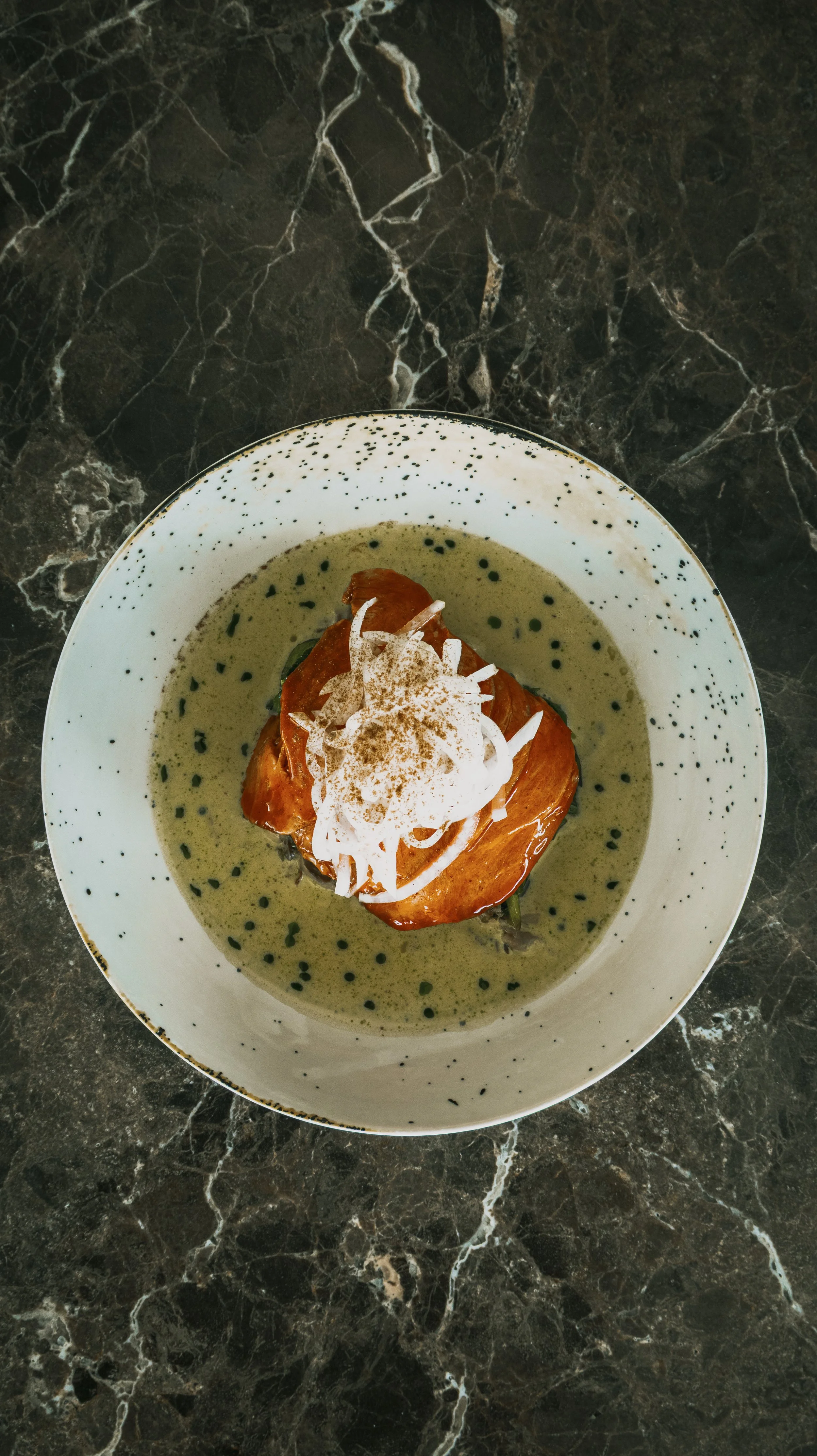 Prato de salmão assado com molho de ervas, coberto com creme de leite, cebola fatiada e uma pitada de especiarias, servido em tigela branca com detalhes pretos, sobre mesa de mármore escuro.
