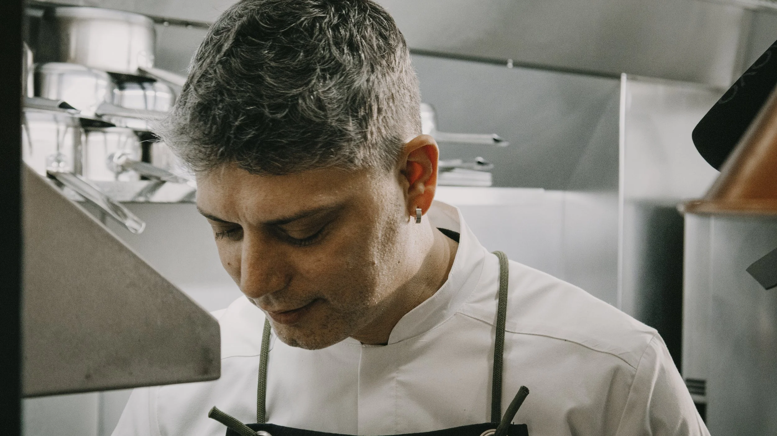 Chef de cozinha com cabelo grisalho e detalhe de piercing na orelha, concentrado, vestindo uniforme branco, no ambiente de cozinha com prateleiras de utensílios metálicos ao fundo.