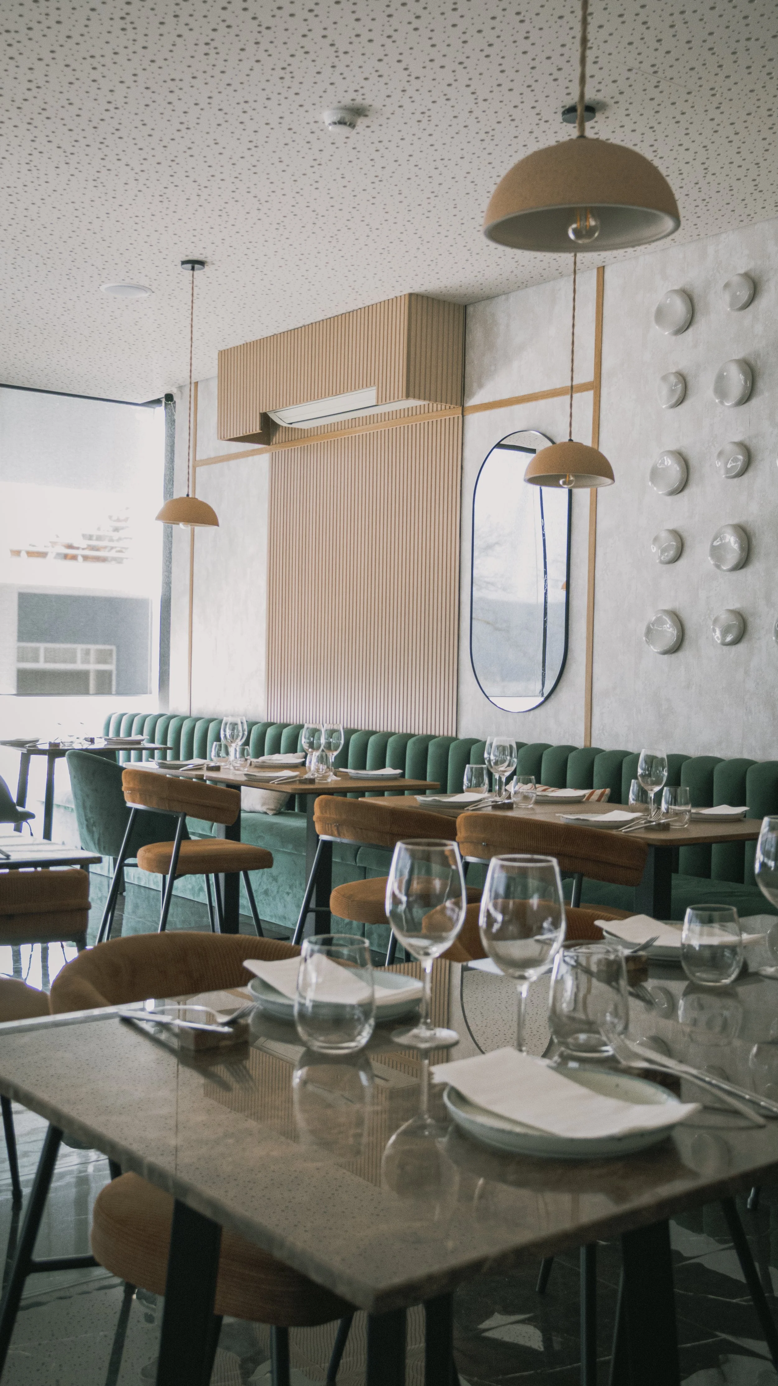 Interior de restaurante com mesa de jantar decorada, copos, pratos e talheres, cadeiras de veludo verde e banco de veludo laranja, parede com decoração de objetos brancos e espelho oval, teto com iluminação suspensa de tom bege.