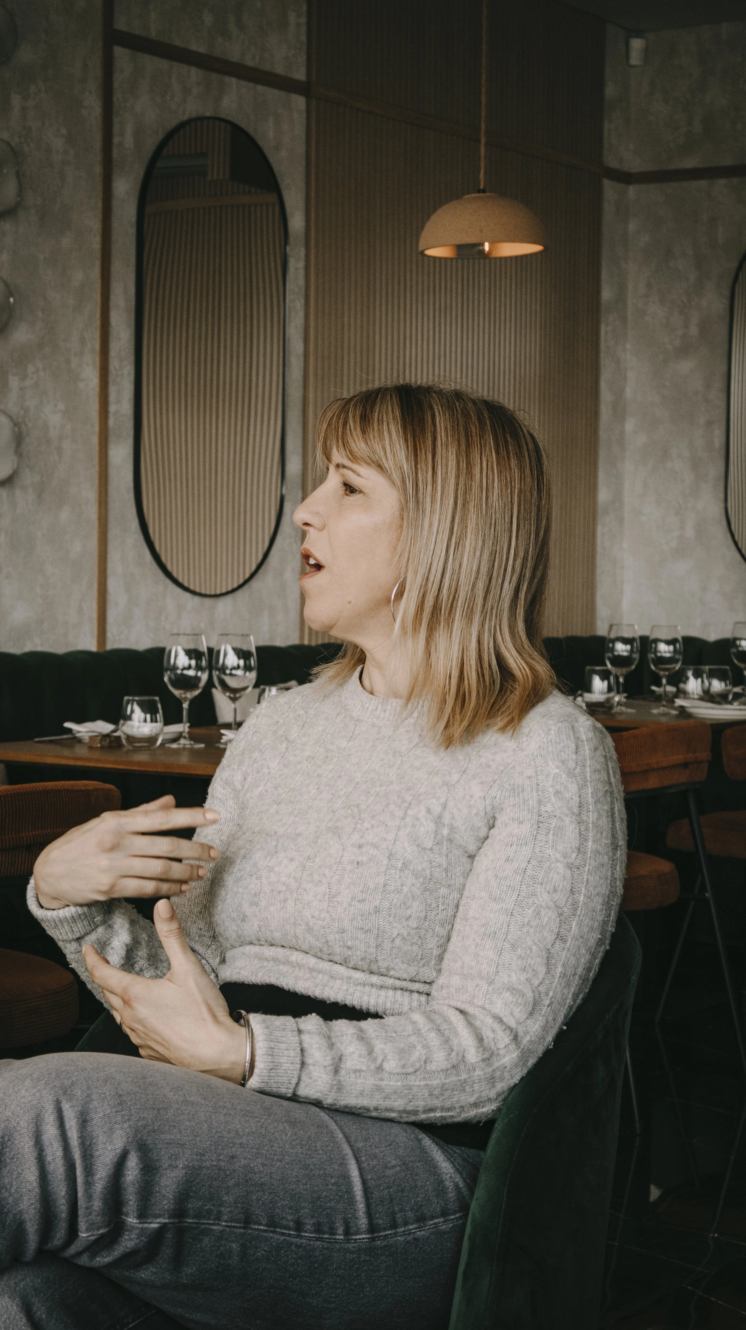 Mulher de cabelo castanho claro, de cabelos médios, usando suéter cinza claro e brincos de argola, sentada em um restaurante, gesticulando com a mão enquanto fala, com decoração moderna e gelo em taças ao fundo.