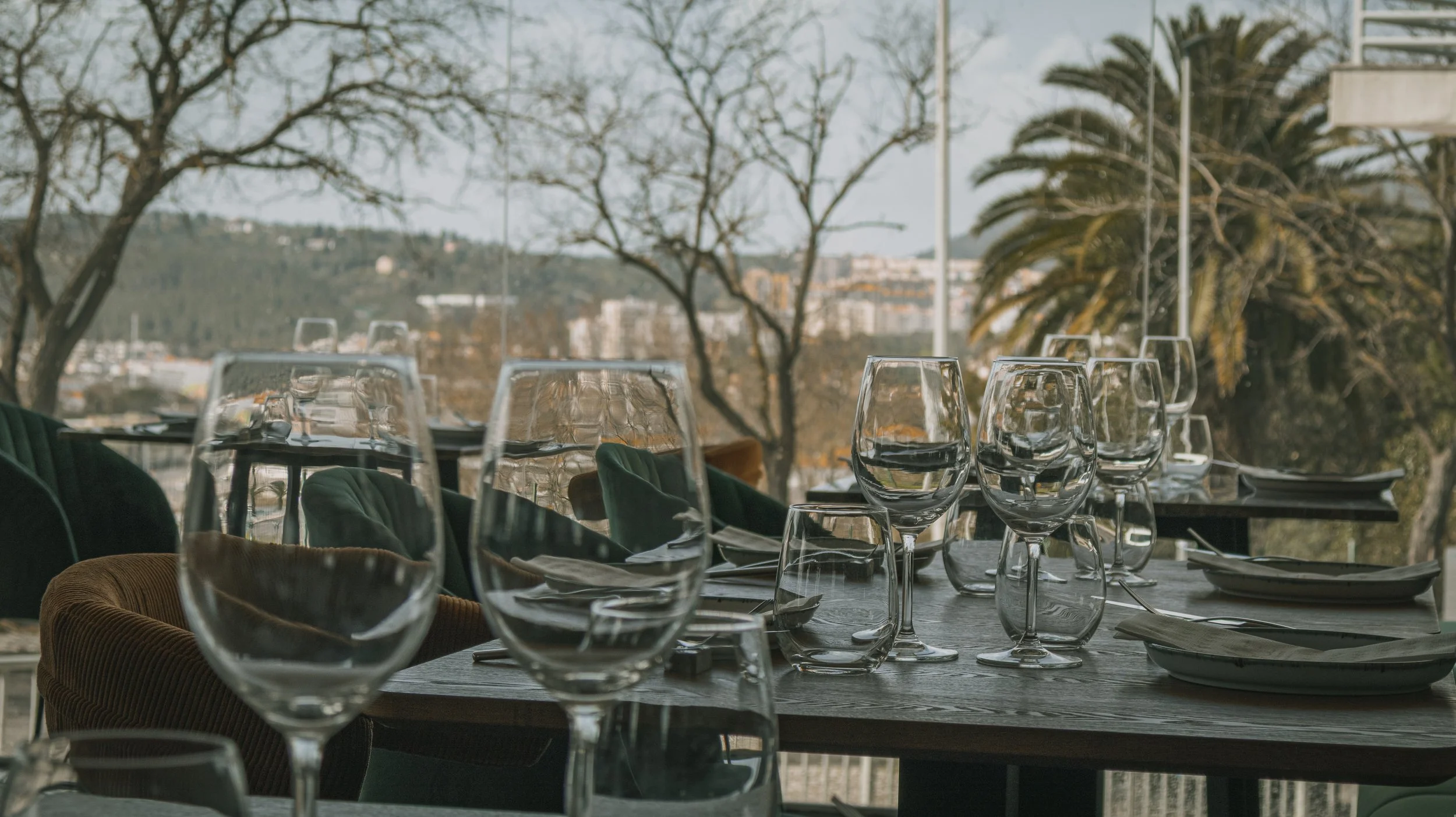 Mesa de jantar elegante ao ar livre com taças de vidro, pratos e talheres, com vista para árvores e cidade ao fundo