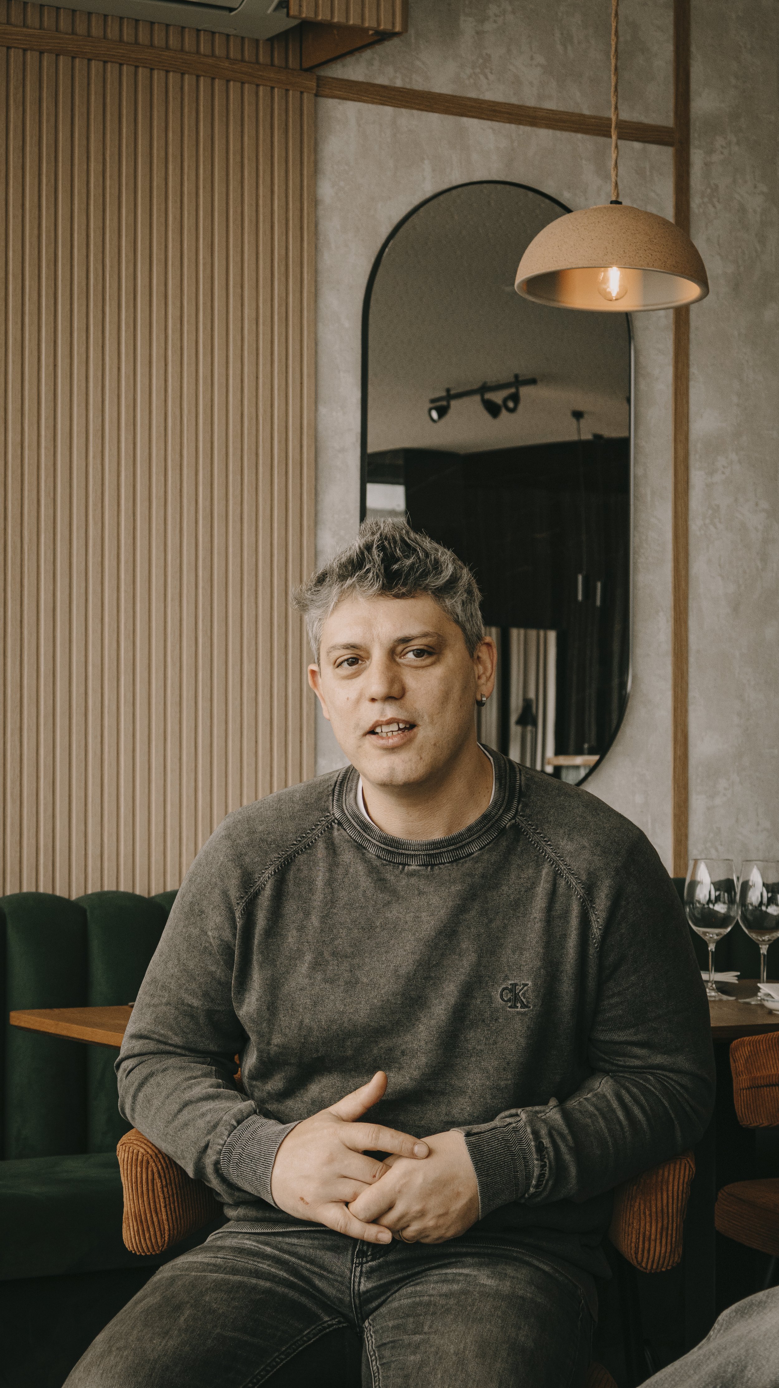 Homem com cabelo curto e gray olhando para a câmera, sentado em uma cadeira ergonômica com almofadas laranja, em um ambiente com decoração moderna, paredes de cimento e madeira, refletido em um espelho oval. Ao fundo, uma luminária suspensa e três ta