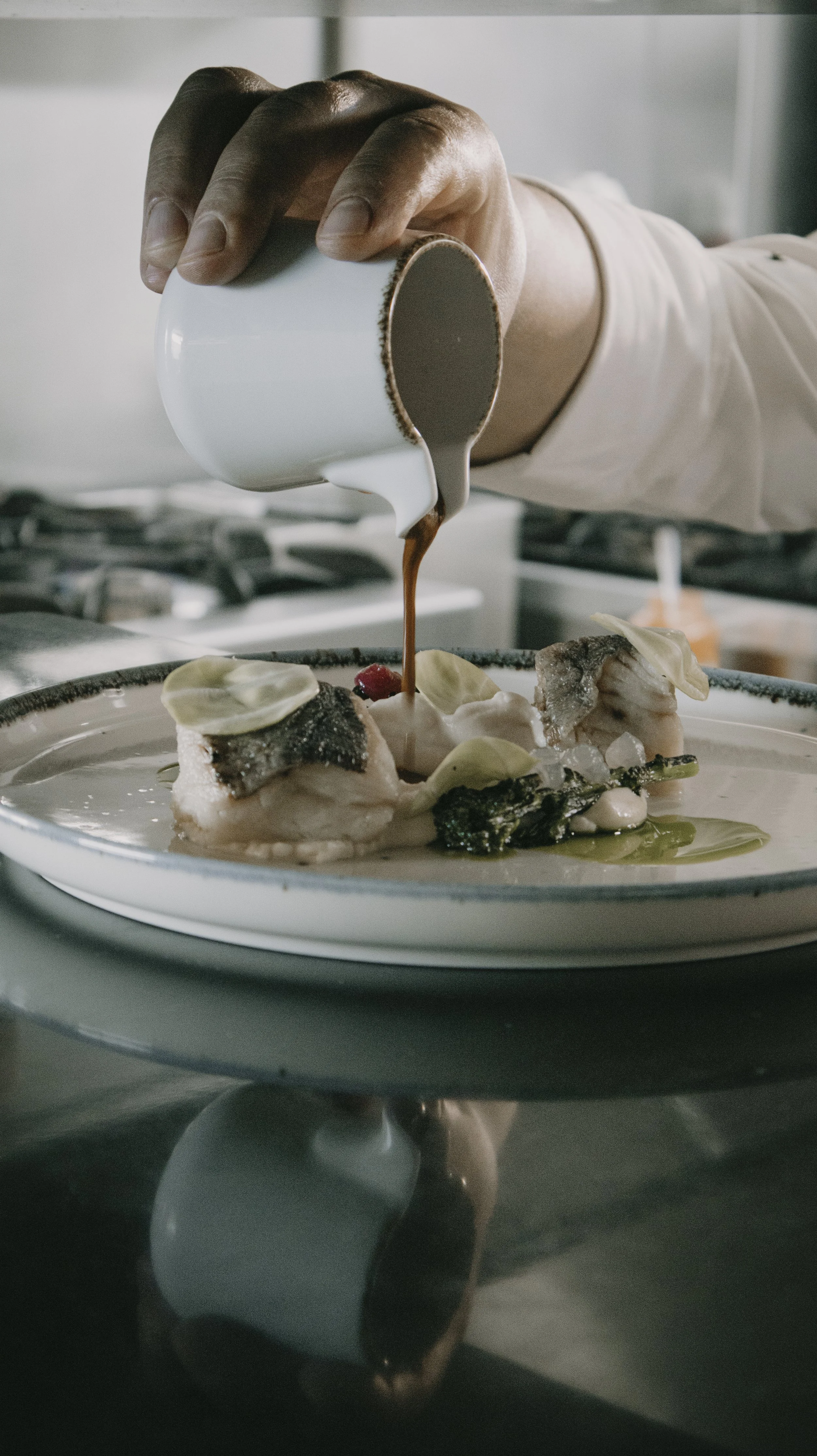 Pessoa despejando molho de chocolate em um prato de sobremesa com peixe, folhas e frutas.