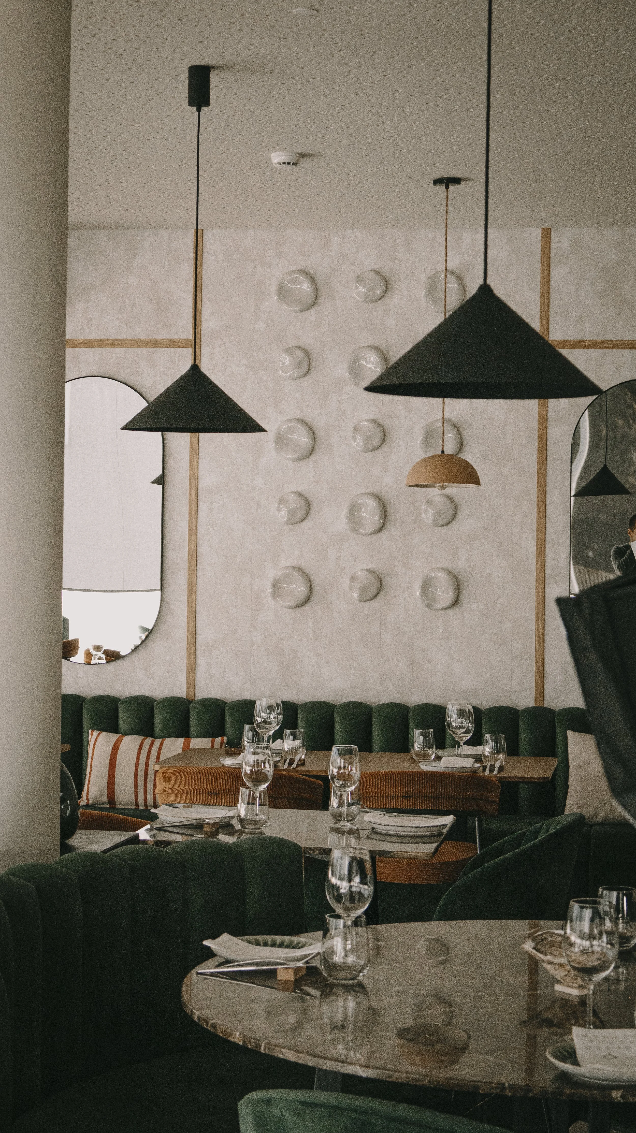 Interior de restaurante com mesas, taças de vidro, parede decorada com pratos e lustres pendurados no teto.