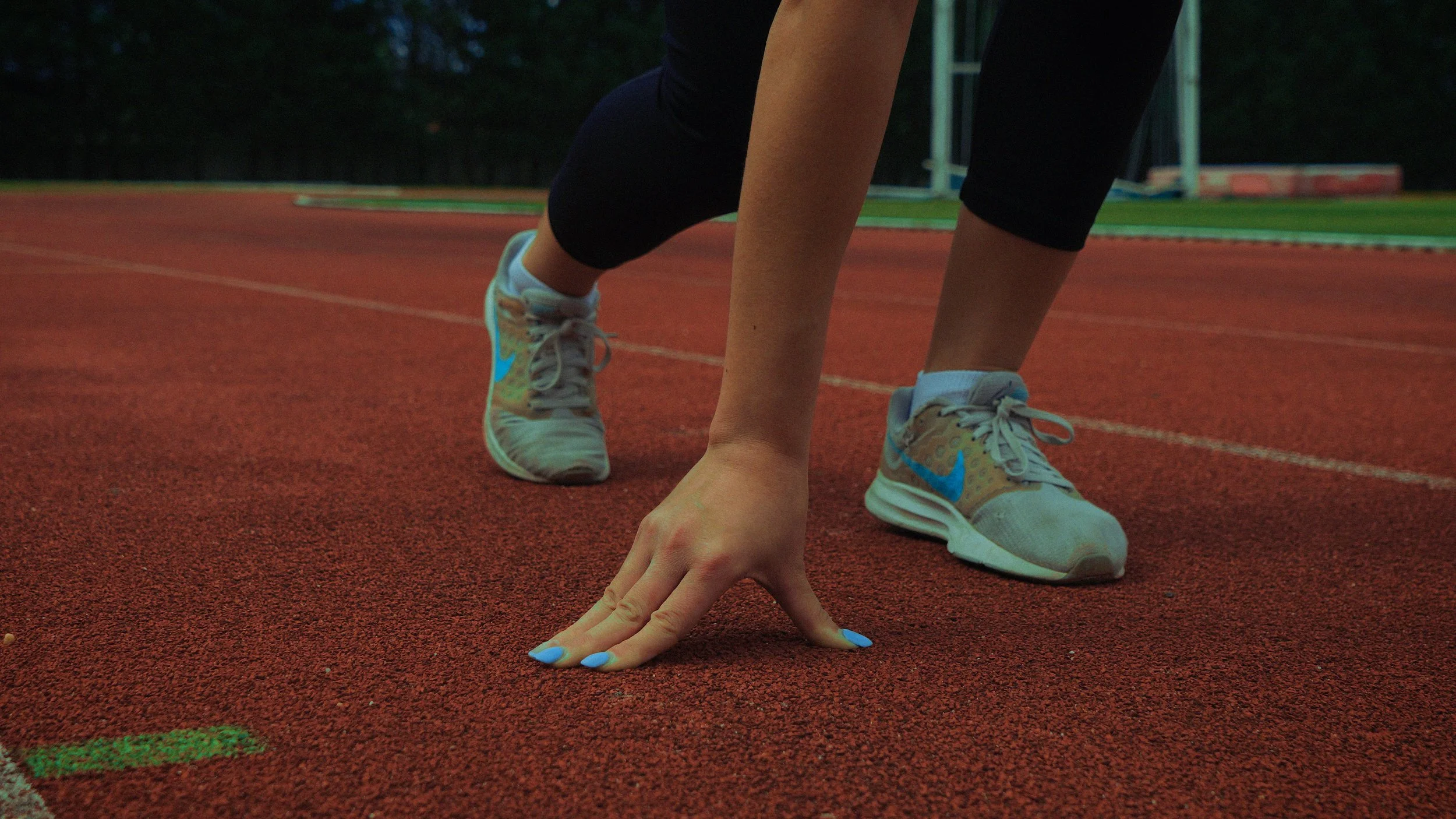 Pessoa agachada na linha de partida de uma pista de atletismo, vestindo tênis esportivos brancos com detalhes azuis e calça preta, com o foco na postura de corrida.