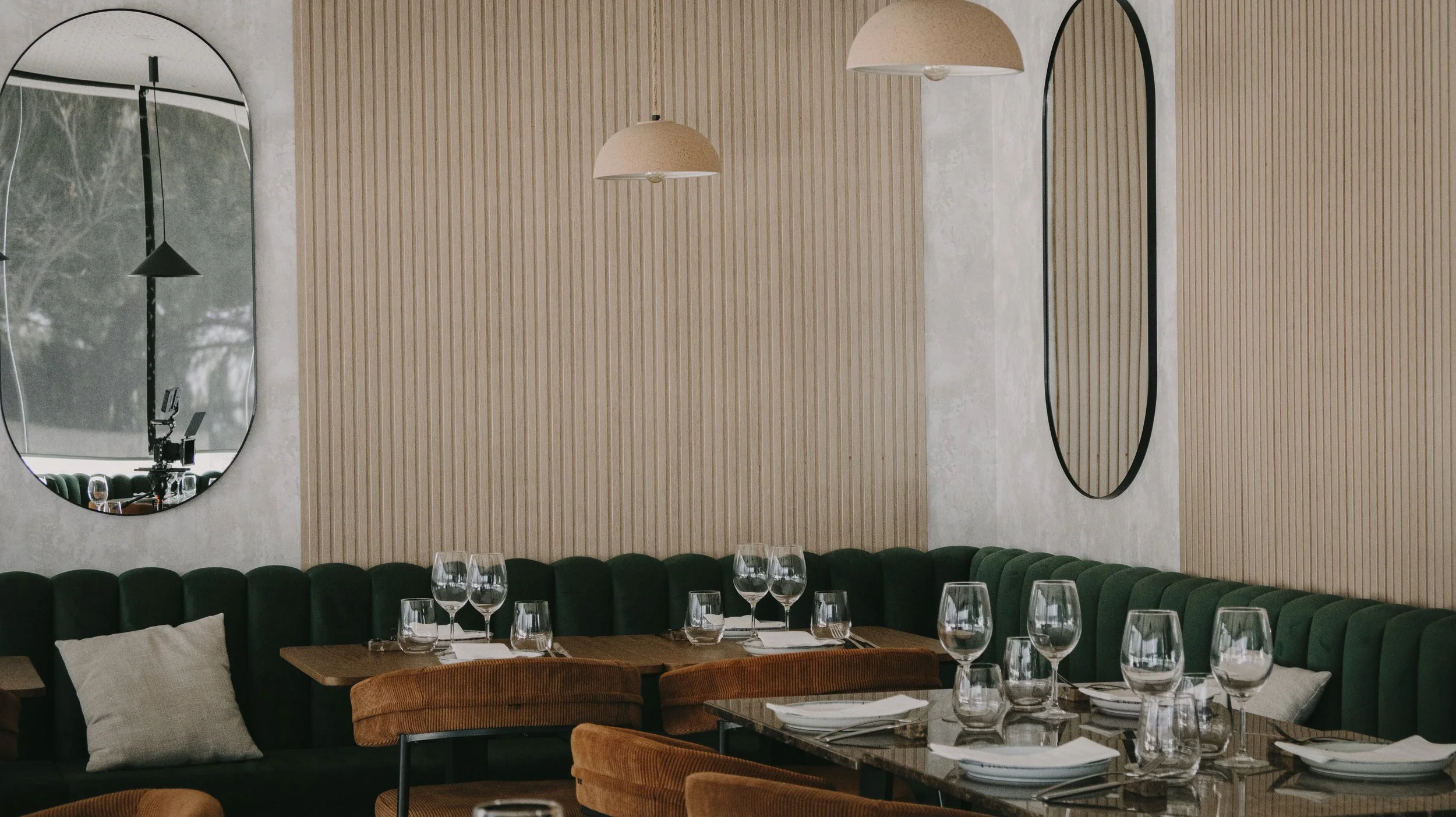 Interior de restaurante com paredes de madeira, espelhos arredondados, poltrona verde, mesa com taças de vinho e talheres, ambiente elegante e moderno