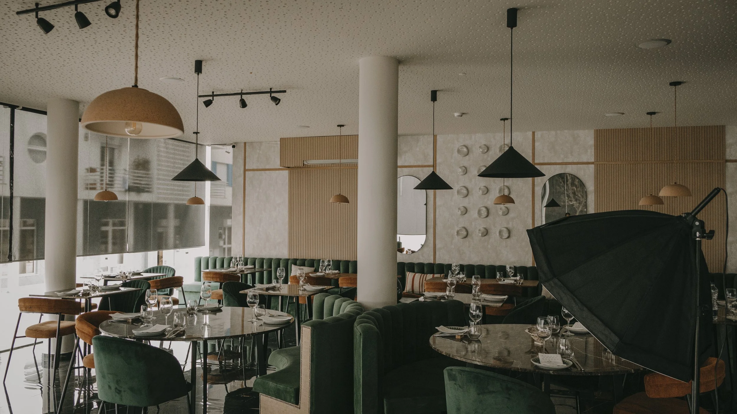 Sala de restaurante elegantemente decorada com mesas, cadeiras e iluminação de pendentes, com uma estrutura moderna e varanda com janelas grandes.