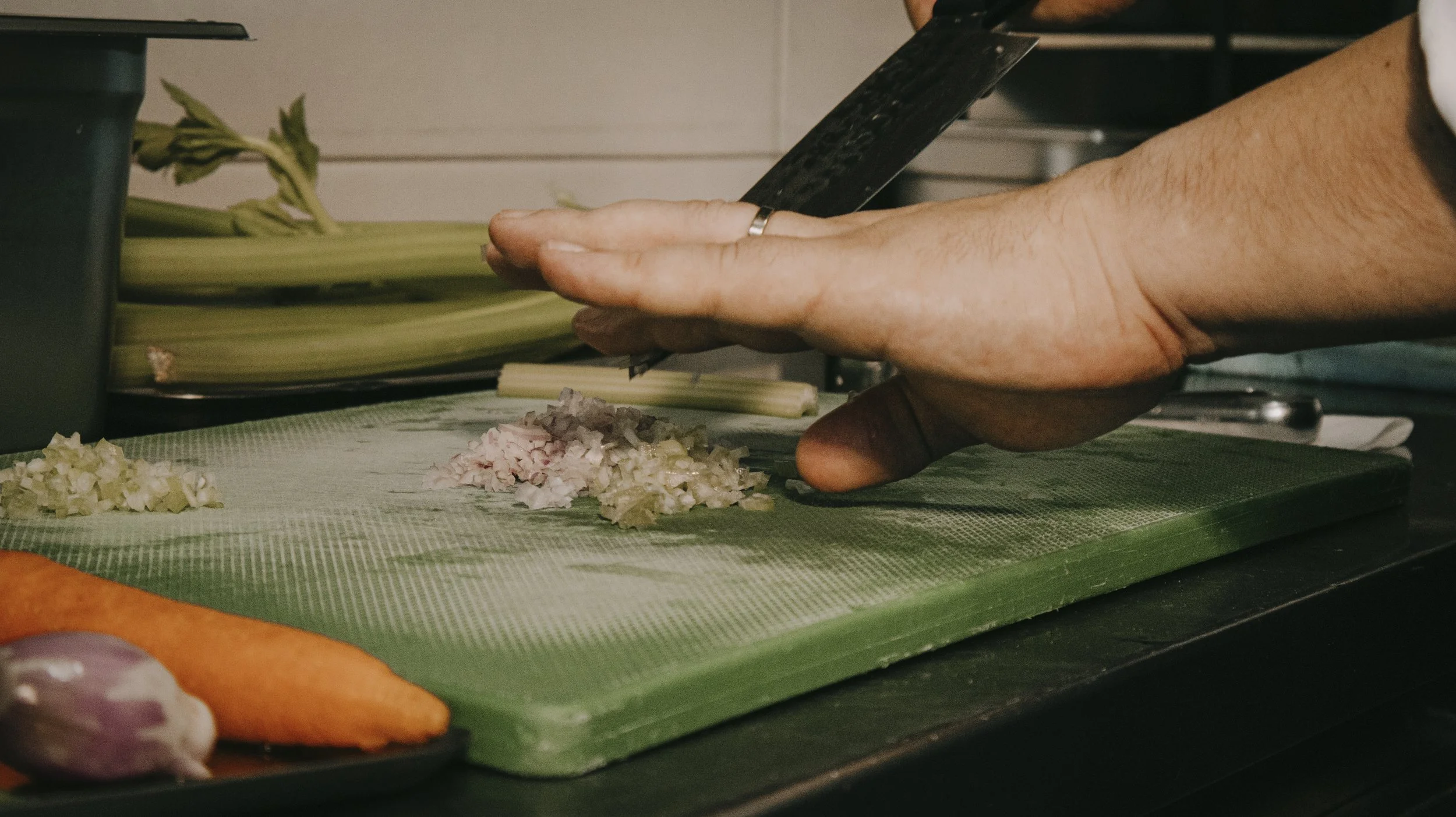 Pessoa picando cebola em uma tábua de corte verde, ao lado de cenoura e alho, em uma cozinha.