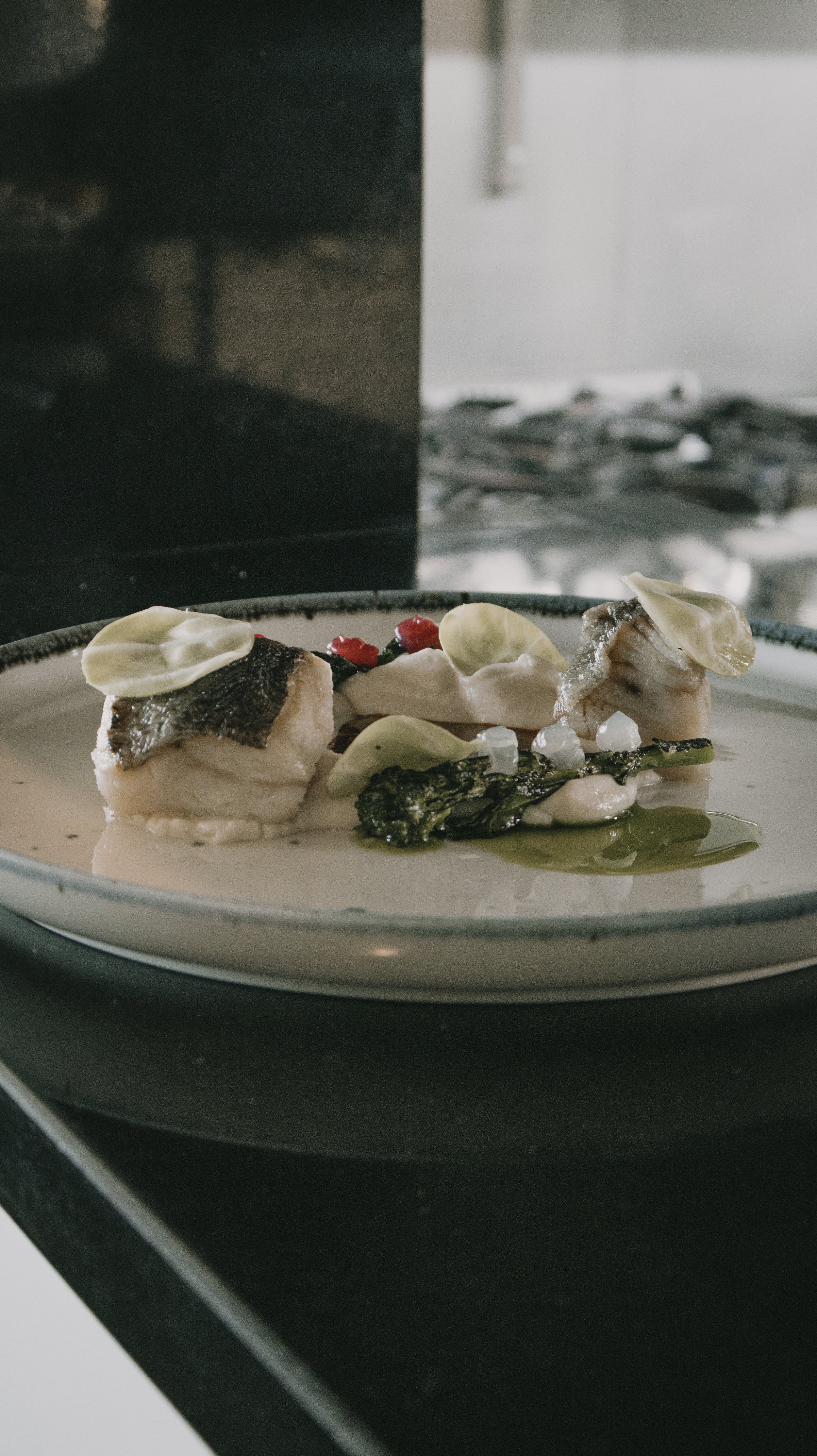 Prato de comida com peixe, verduras grelhadas, e molho, decorado com folhas verdes e pequenas flores comestíveis.
