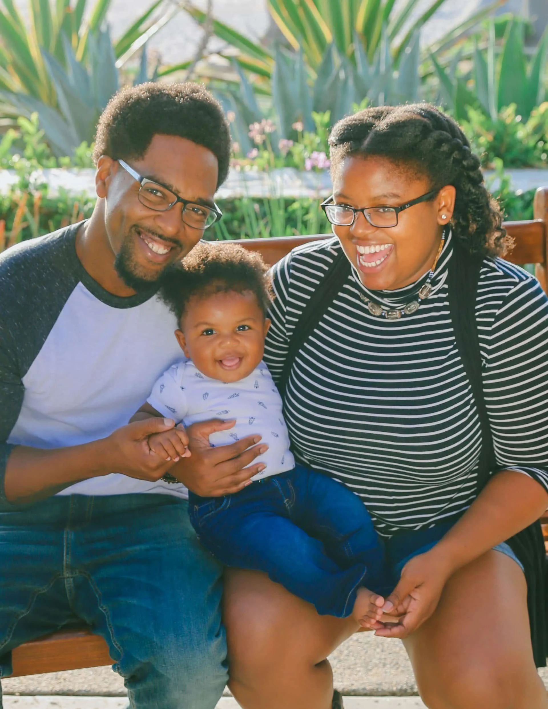 Smiling Black couple sitting on a bench outdoors, holding a happy baby between them, with green plants in the background.