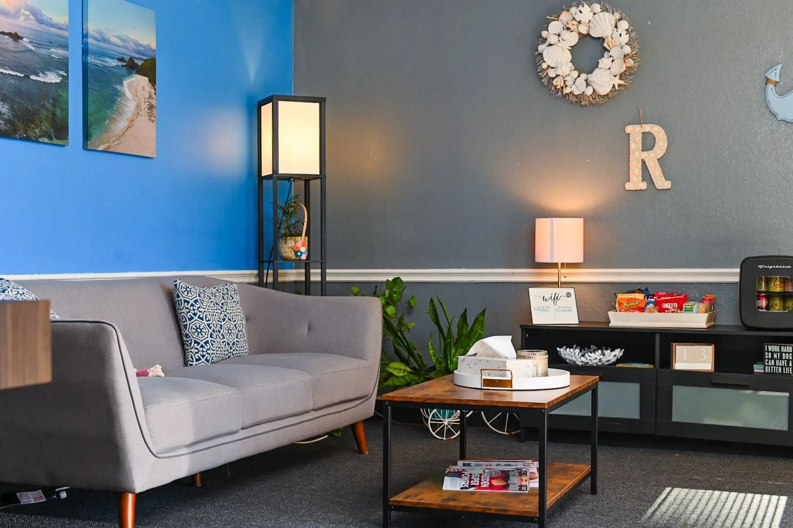 A cozy waiting room with a gray sofa, coffee table, snacks, plants, and wall art on blue and gray walls.