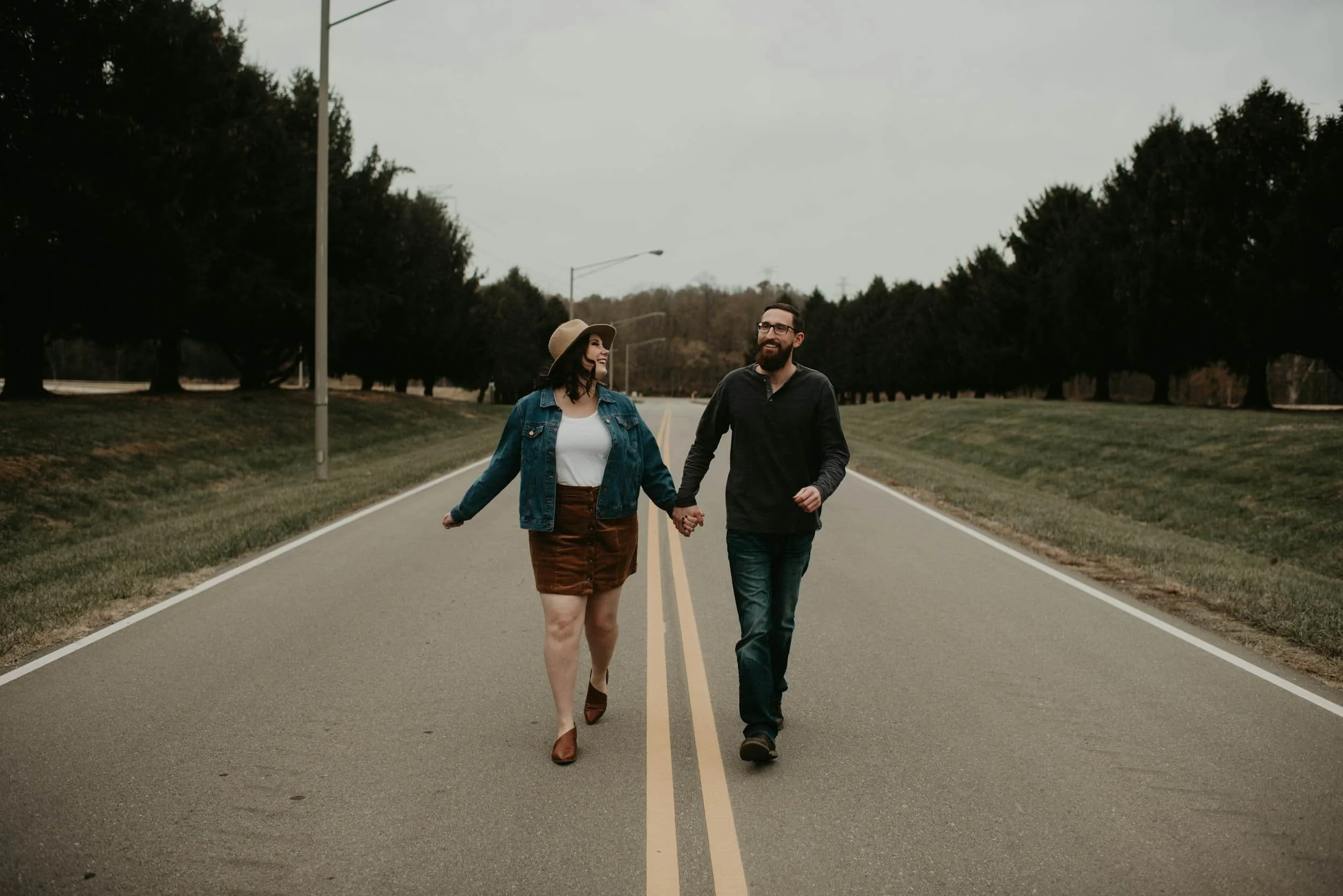 A couple holding hands and walking down the middle of an empty road lined with trees on a cloudy day.