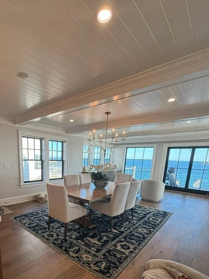 Modern dining room with a wooden table, six cream-colored upholstered chairs, a black and white patterned rug, large windows overlooking water, and a chandelier with candles.