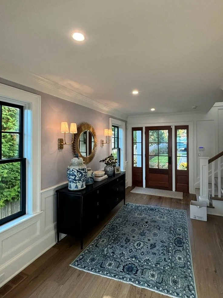 Entryway with a dark wood sideboard, decorative vases, mirror, and wall sconces, with windows and a front door.