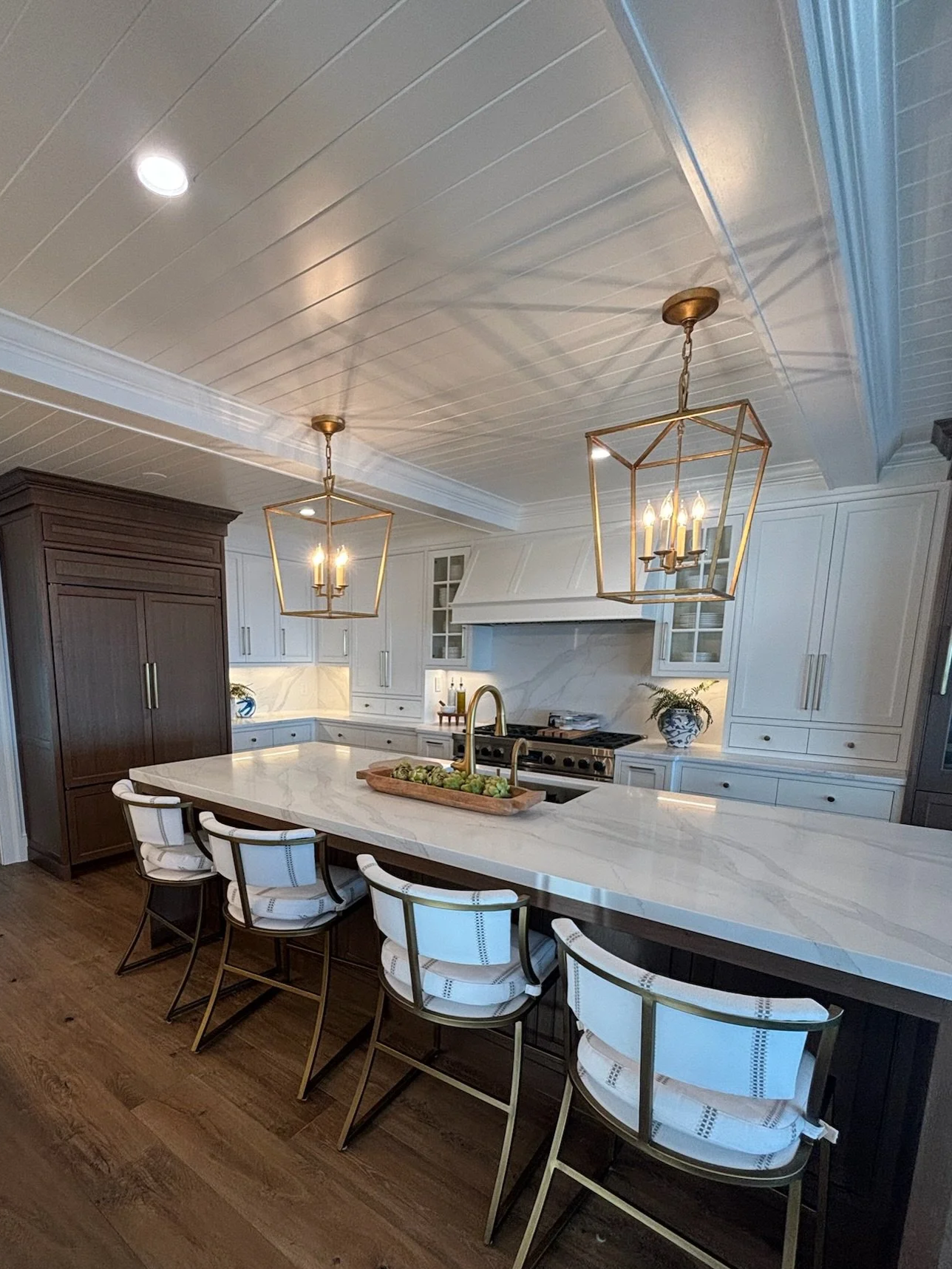 A modern kitchen with a large white marble island, dark brown and white cabinetry, gold pendant lights, and wooden flooring.
