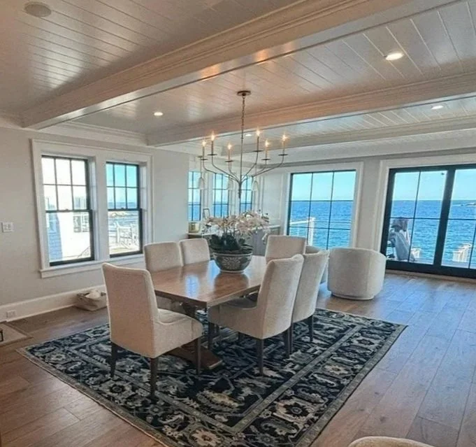 Dining room with a wooden table, eight upholstered chairs, a floral centerpiece, a chandelier, large windows and sliding glass doors with views of the water.