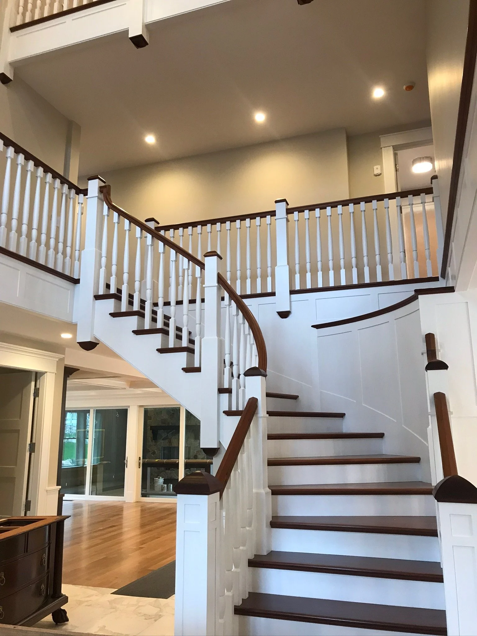 Interior view of a house featuring a staircase with white railings, dark wood handrails, and steps. The space has recessed ceiling lights and a partially visible sliding glass door leading to an outdoor area with a brick fireplace.