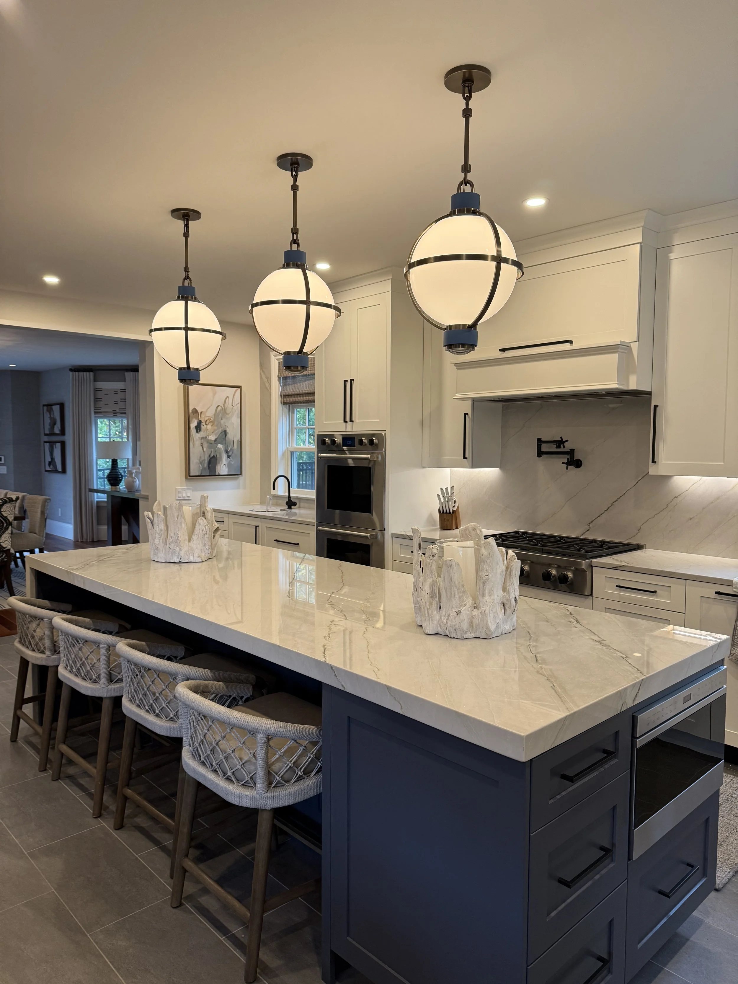 Modern kitchen with white cabinets, marble backsplash, and a large blue island with six upholstered bar stools. Three pendant lights hang over the island.